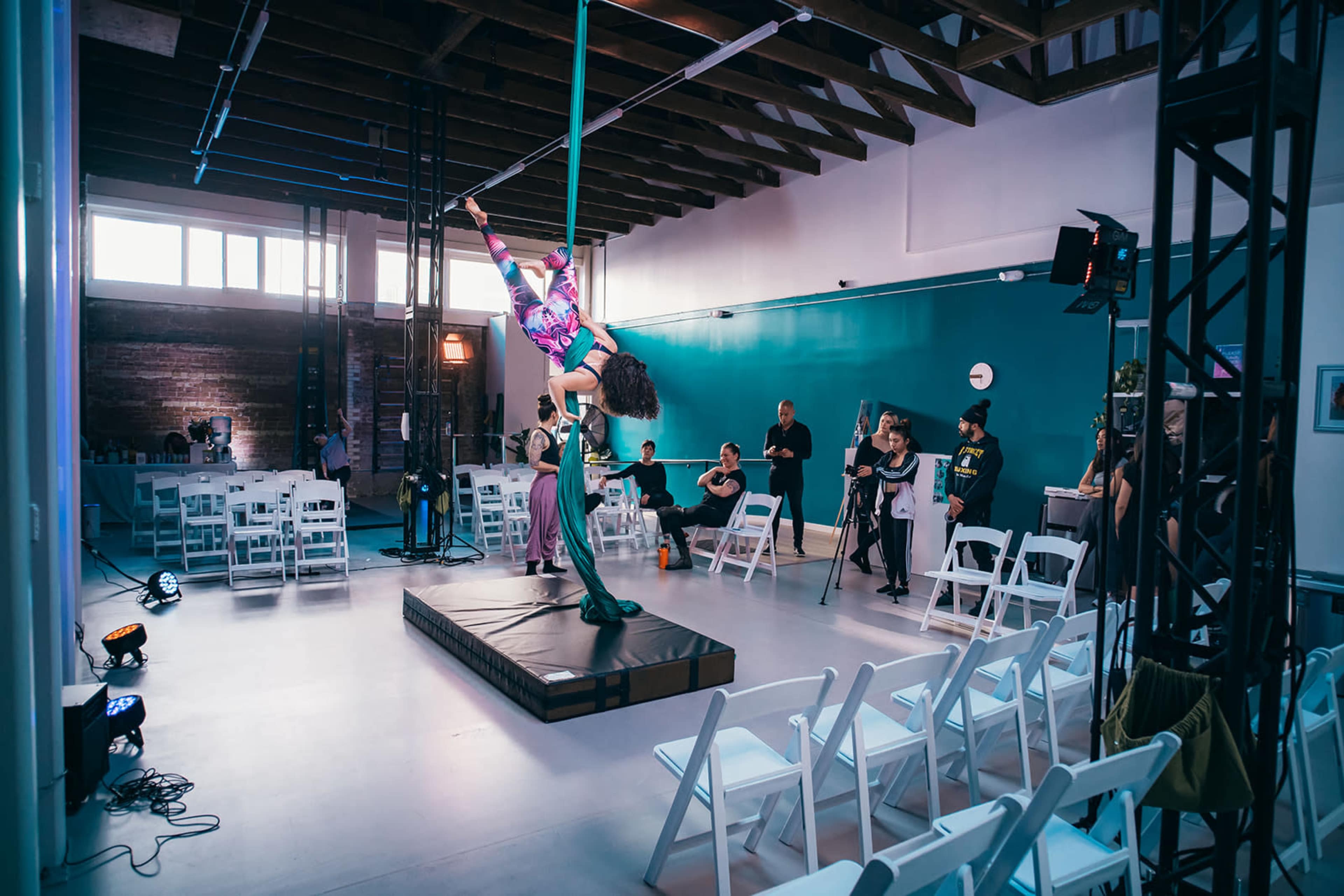 A performer demonstrates aerial skills on a pole in a spacious venue with an audience seated in white chairs.