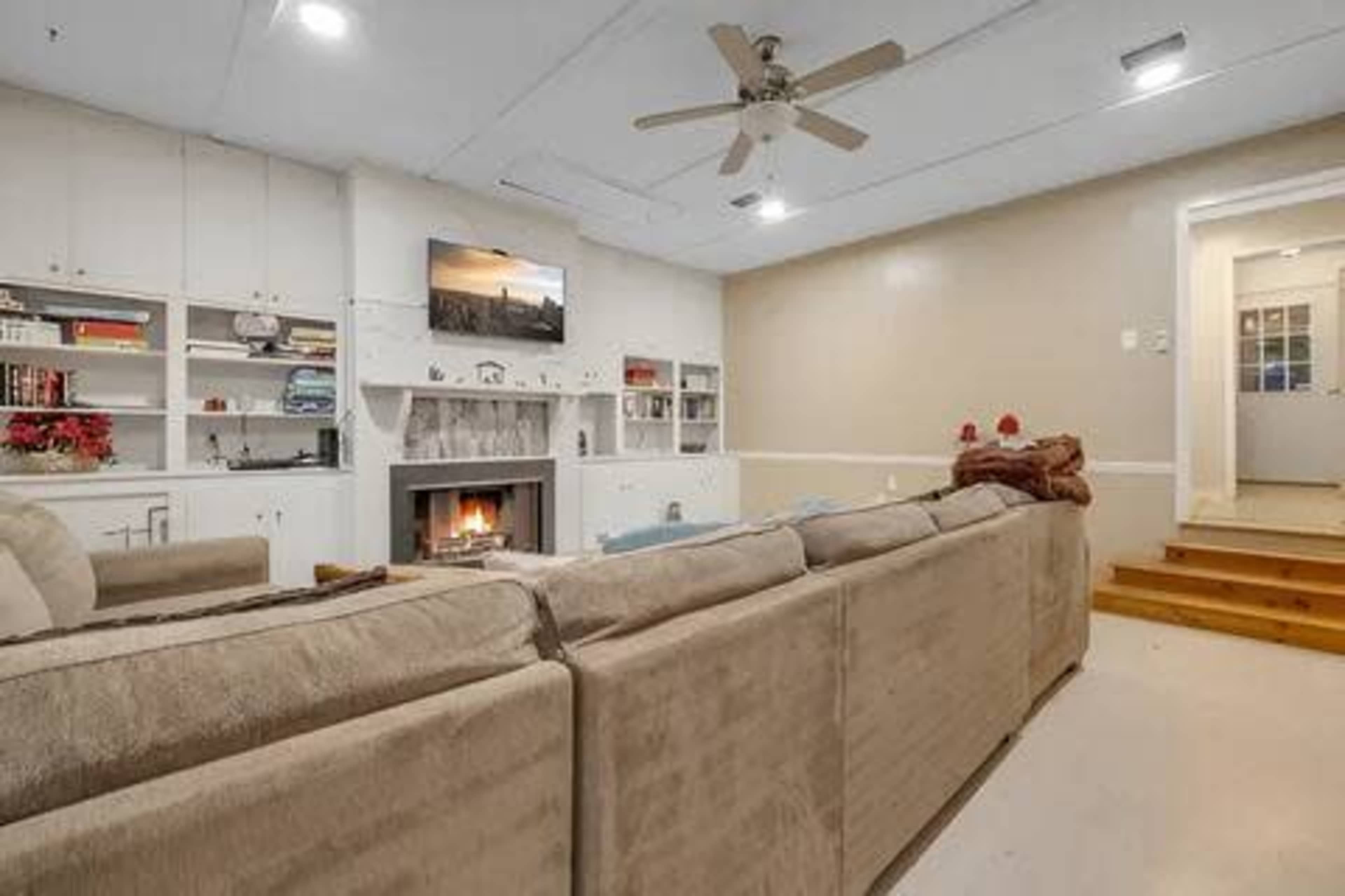 A living room with a large sectional sofa facing a fireplace and television, with built-in shelves and a staircase leading to another area in the background.