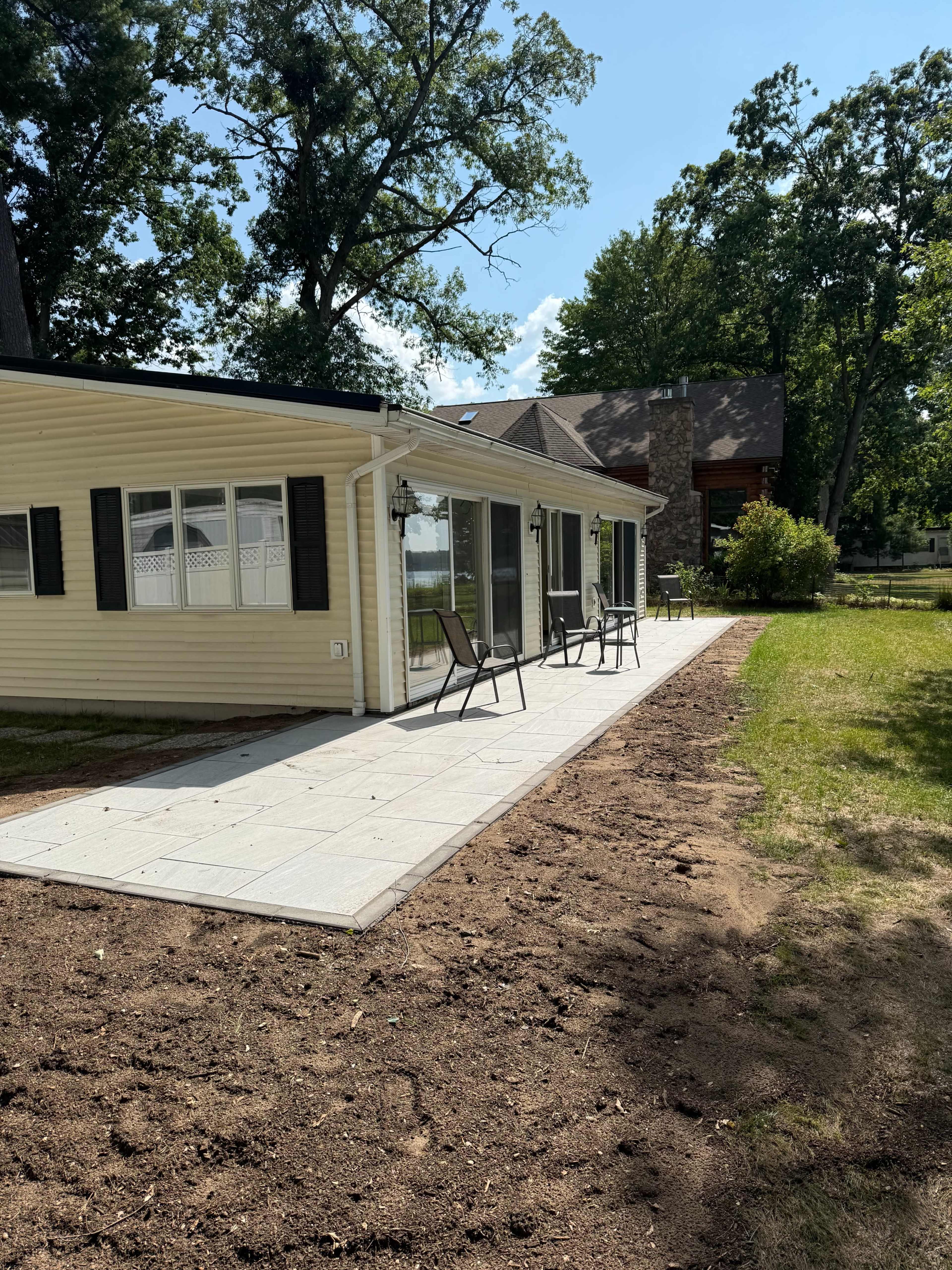 The image shows a yellow house with a patio made of square tiles, accompanied by several chairs, and a lawn area that has been recently disturbed or prepared for planting.