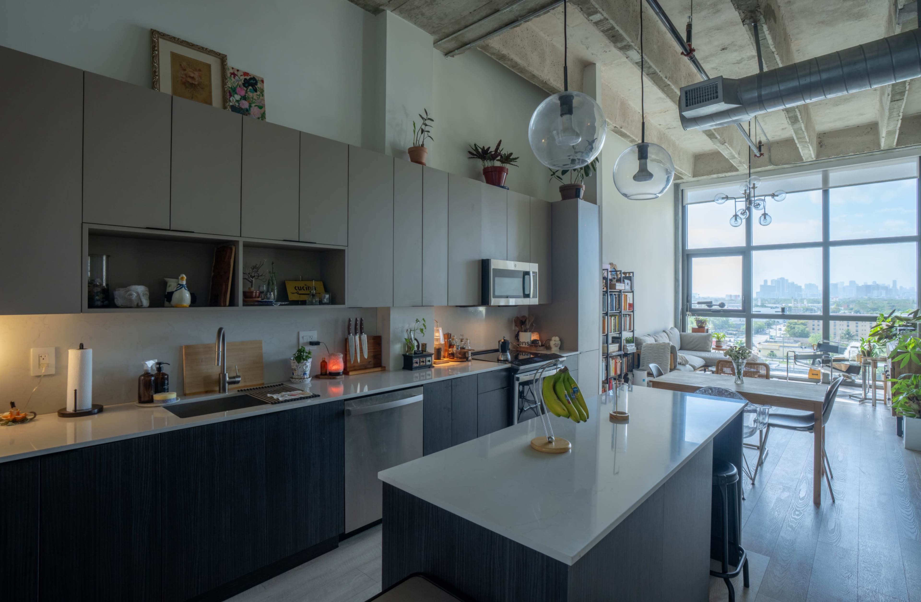 The image shows a modern kitchen with sleek cabinets, a central island, and a large window that offers a view of a city skyline.