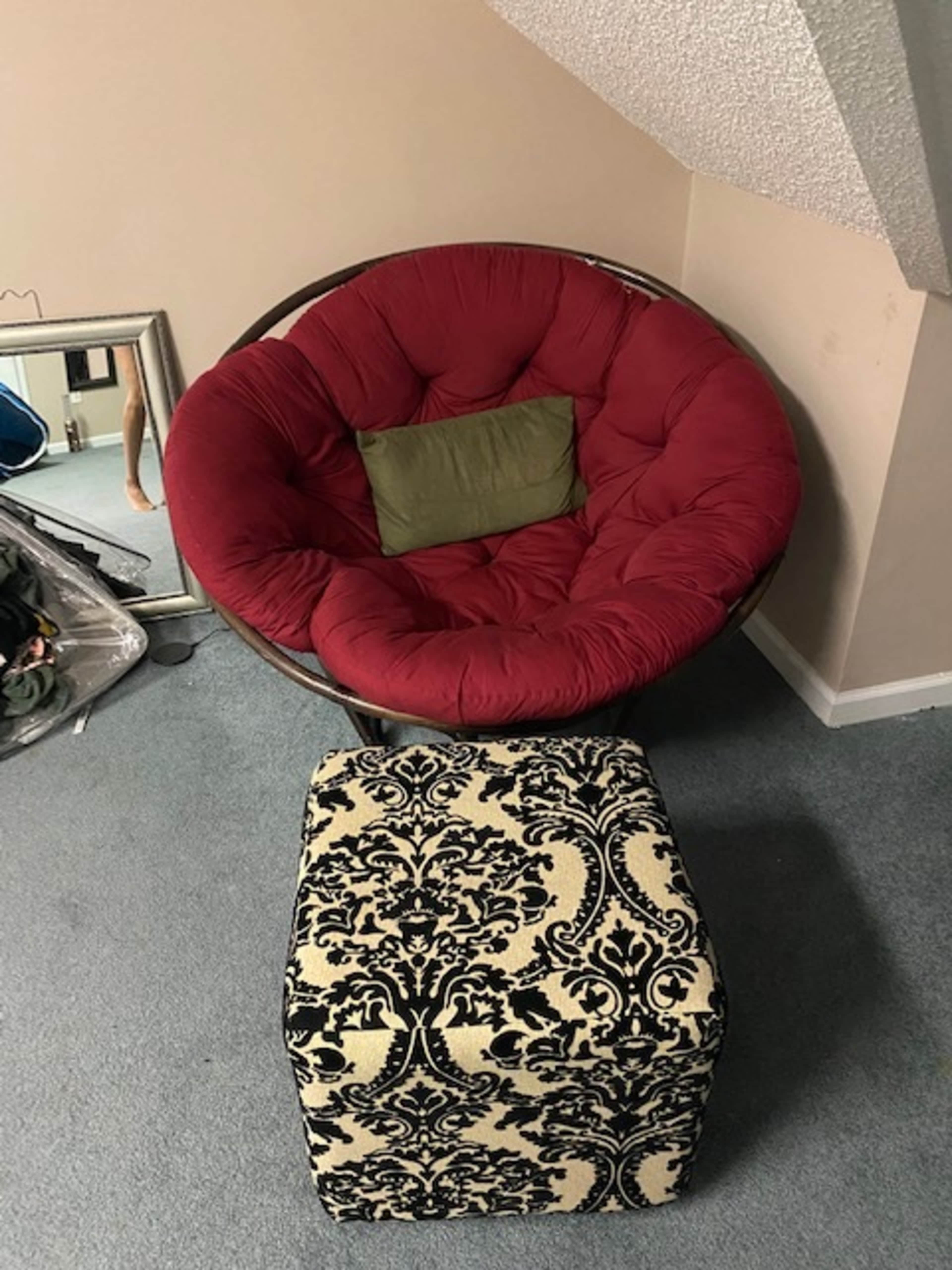 The image shows a red, circular cushioned chair paired with a black and cream patterned ottoman, situated on a carpeted floor in a room.