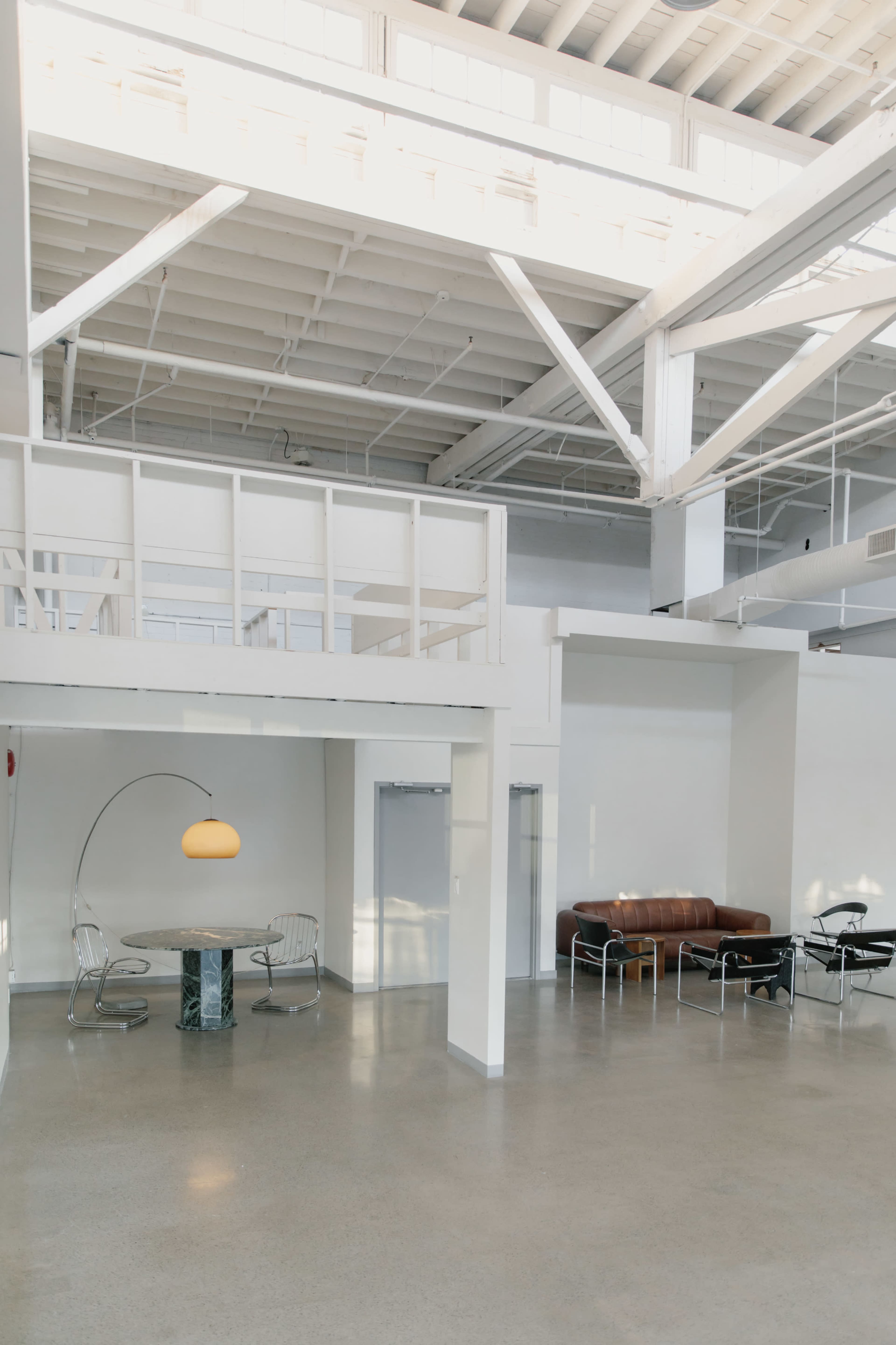 The image shows a spacious, modern interior featuring a table with a floor lamp, a brown leather sofa, and various chairs in a minimalist design.