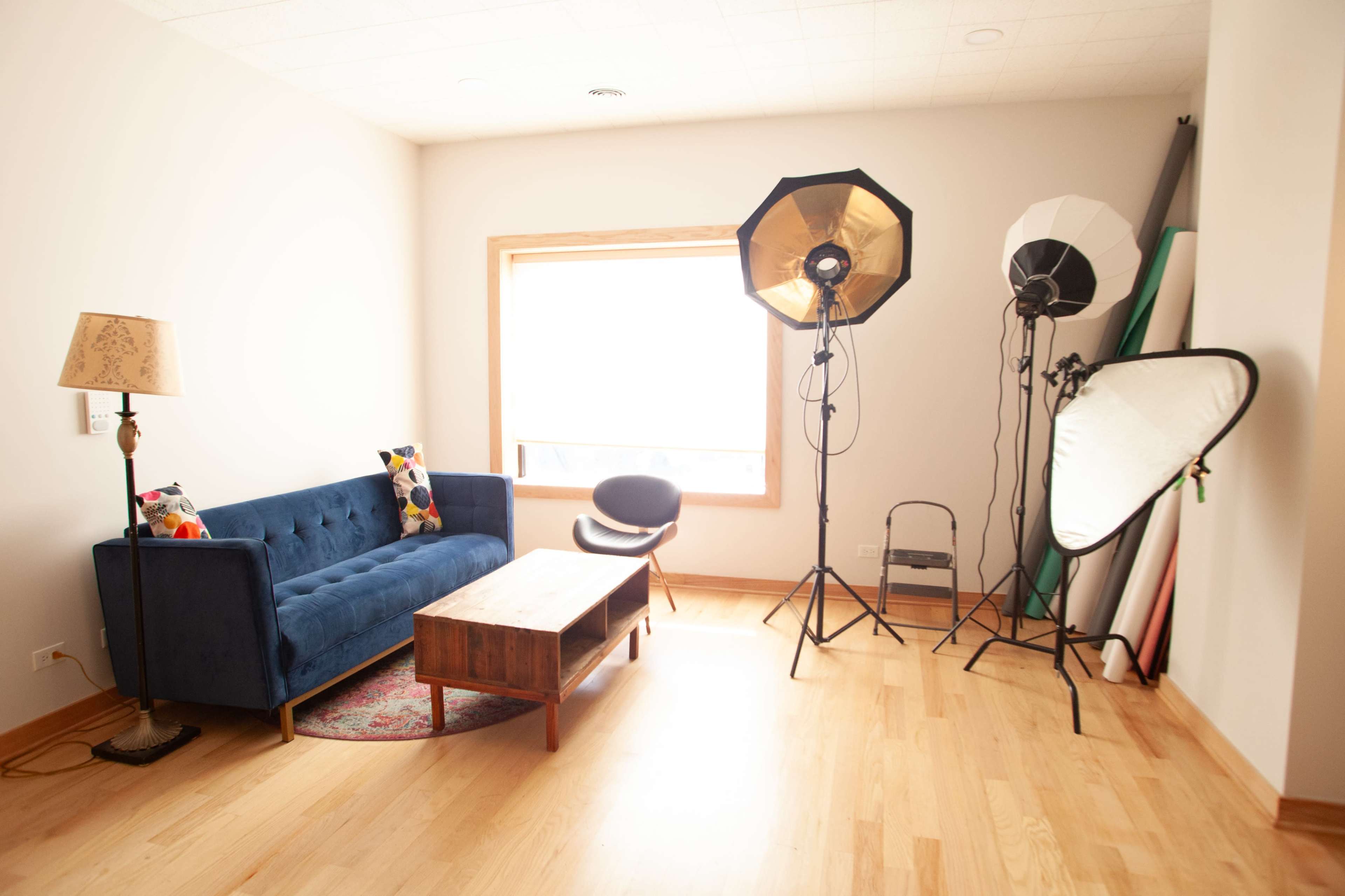 A cozy living room features a blue sofa, a wooden coffee table, and studio lighting equipment next to a large window.