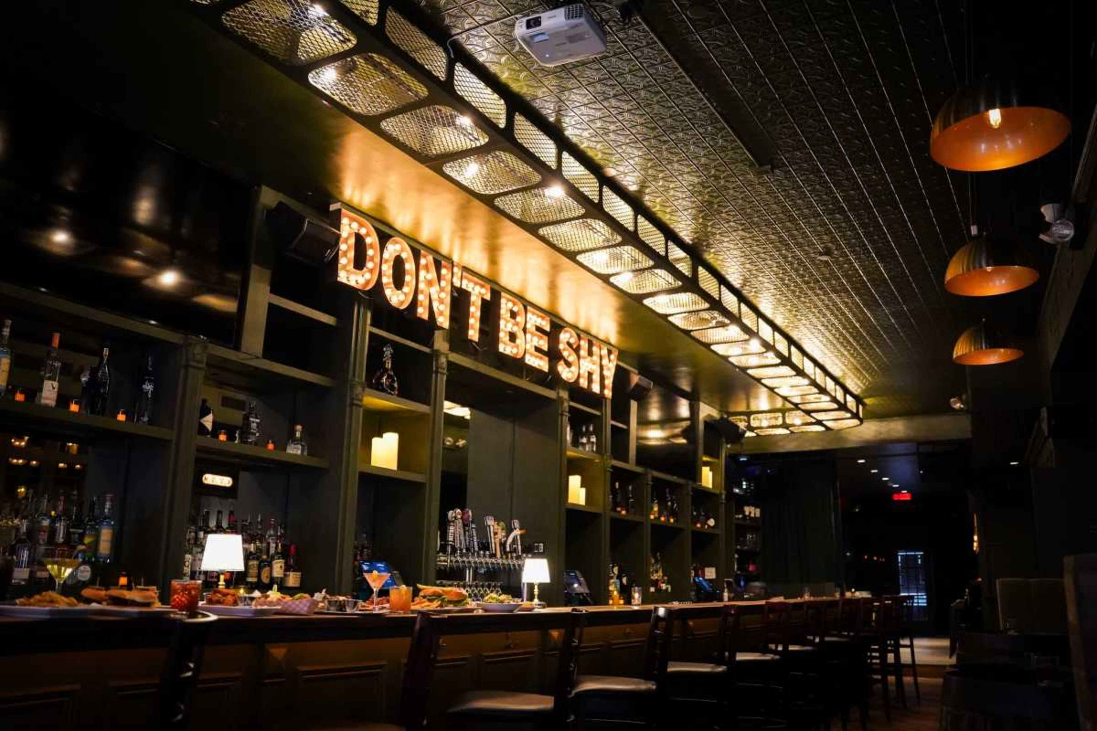 The image shows a dimly lit bar with a long countertop, shelves of liquor, and a neon sign that reads "DON'T BE SHY" illuminated above the bar.