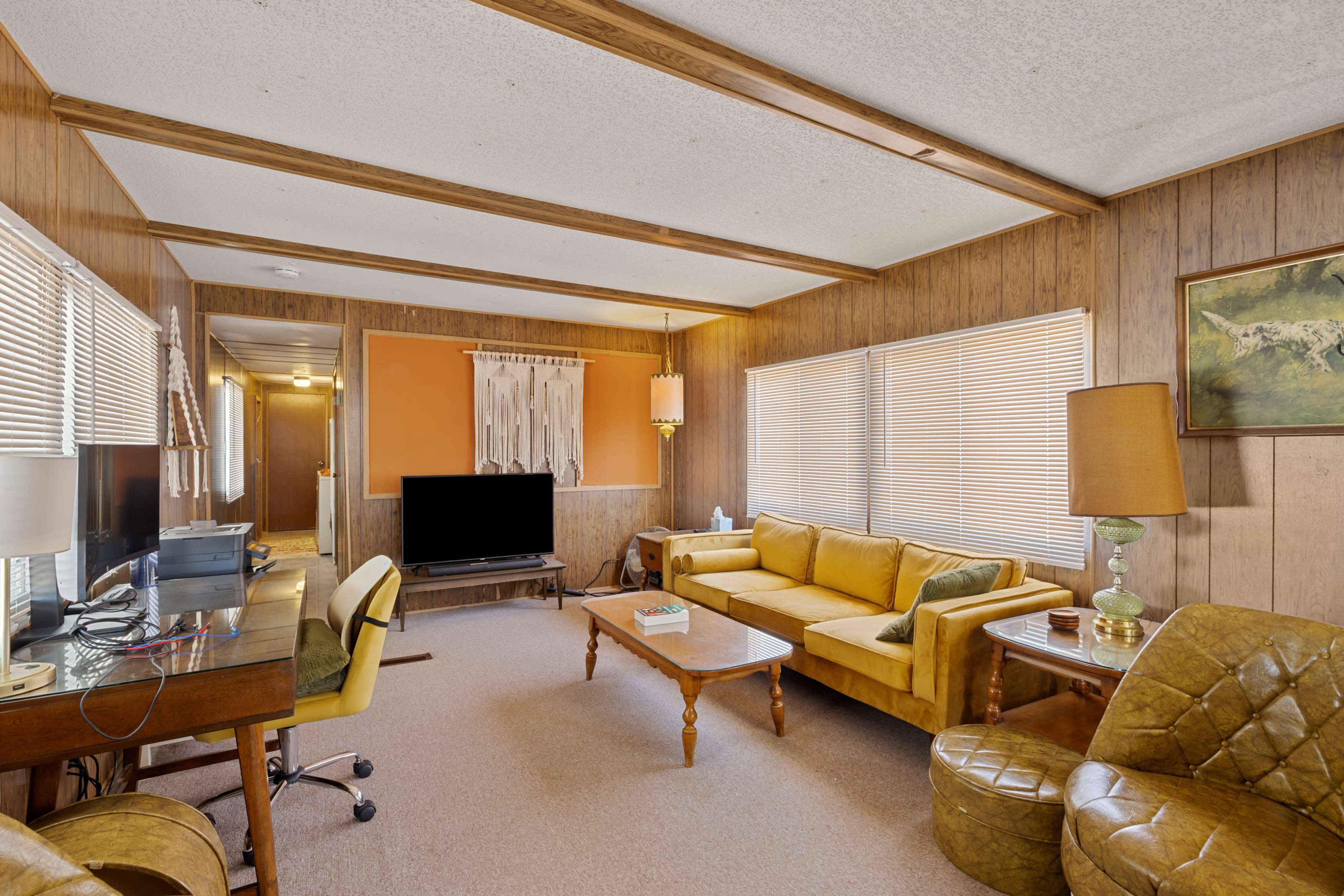 The image shows a retro-style living room featuring wood-paneled walls, a yellow sofa, a coffee table, a TV, and a desk with a chair.