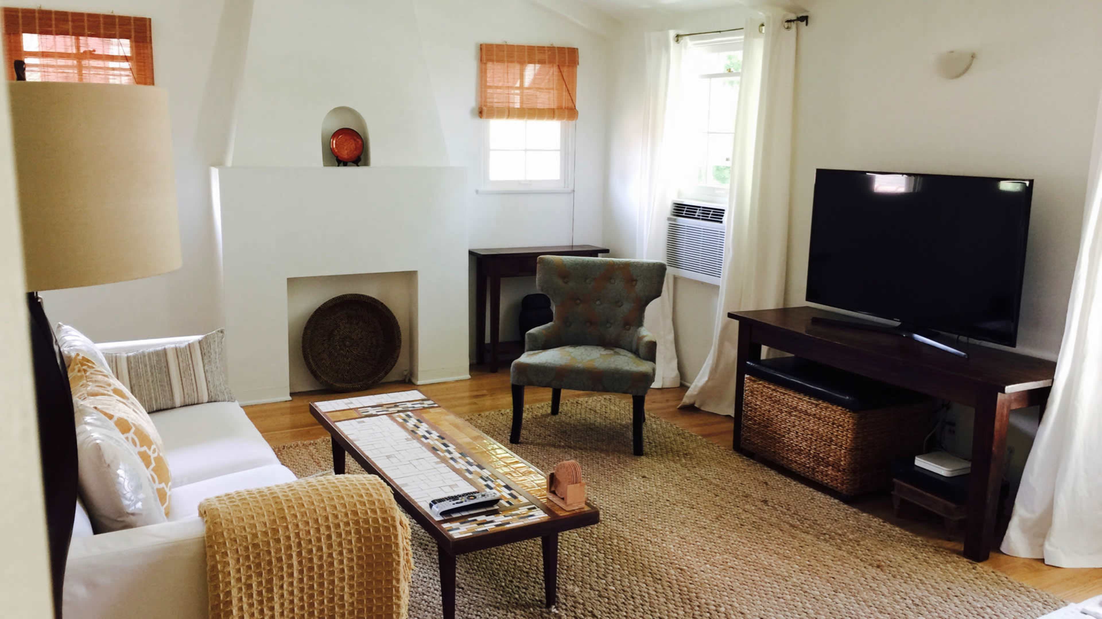 The living room features a white sofa, a gray upholstered chair, a wooden coffee table, and a large TV, all arranged around a woven area rug.