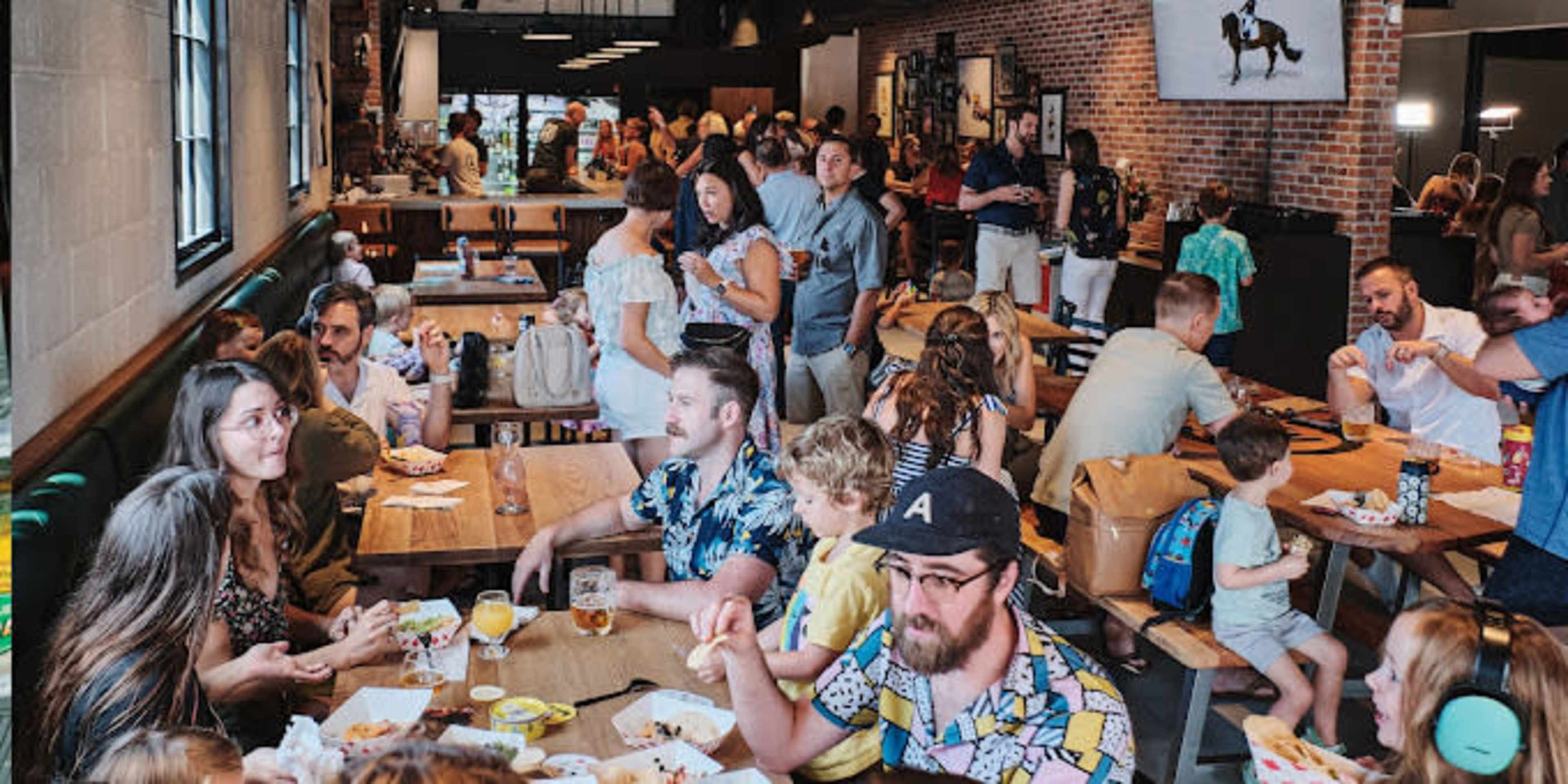A bustling restaurant scene shows groups of people eating and socializing at wooden tables, with a mix of children and adults throughout the space.