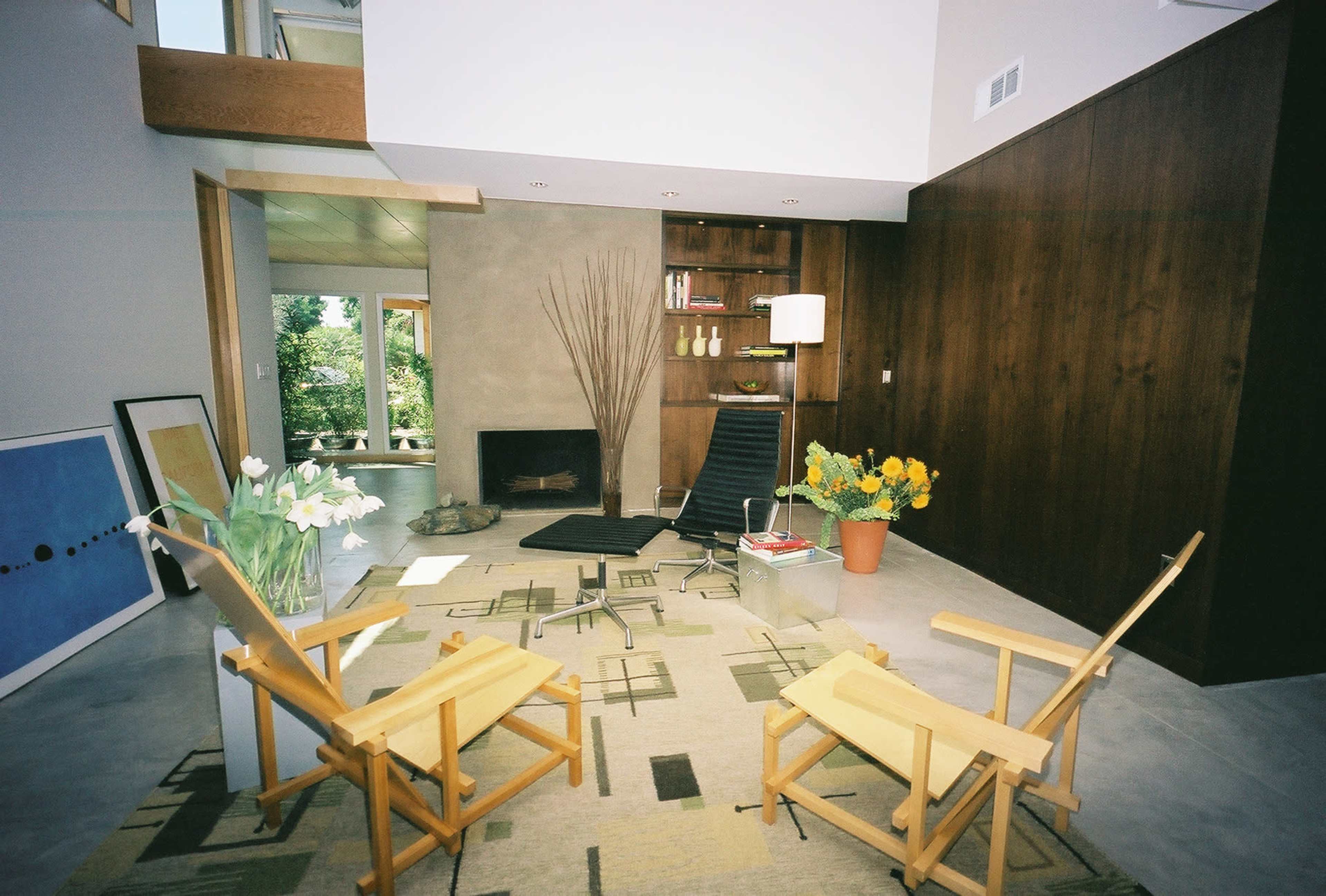 A modern living room with two wooden chairs facing a coffee table on a patterned rug, surrounded by plants and a fireplace.