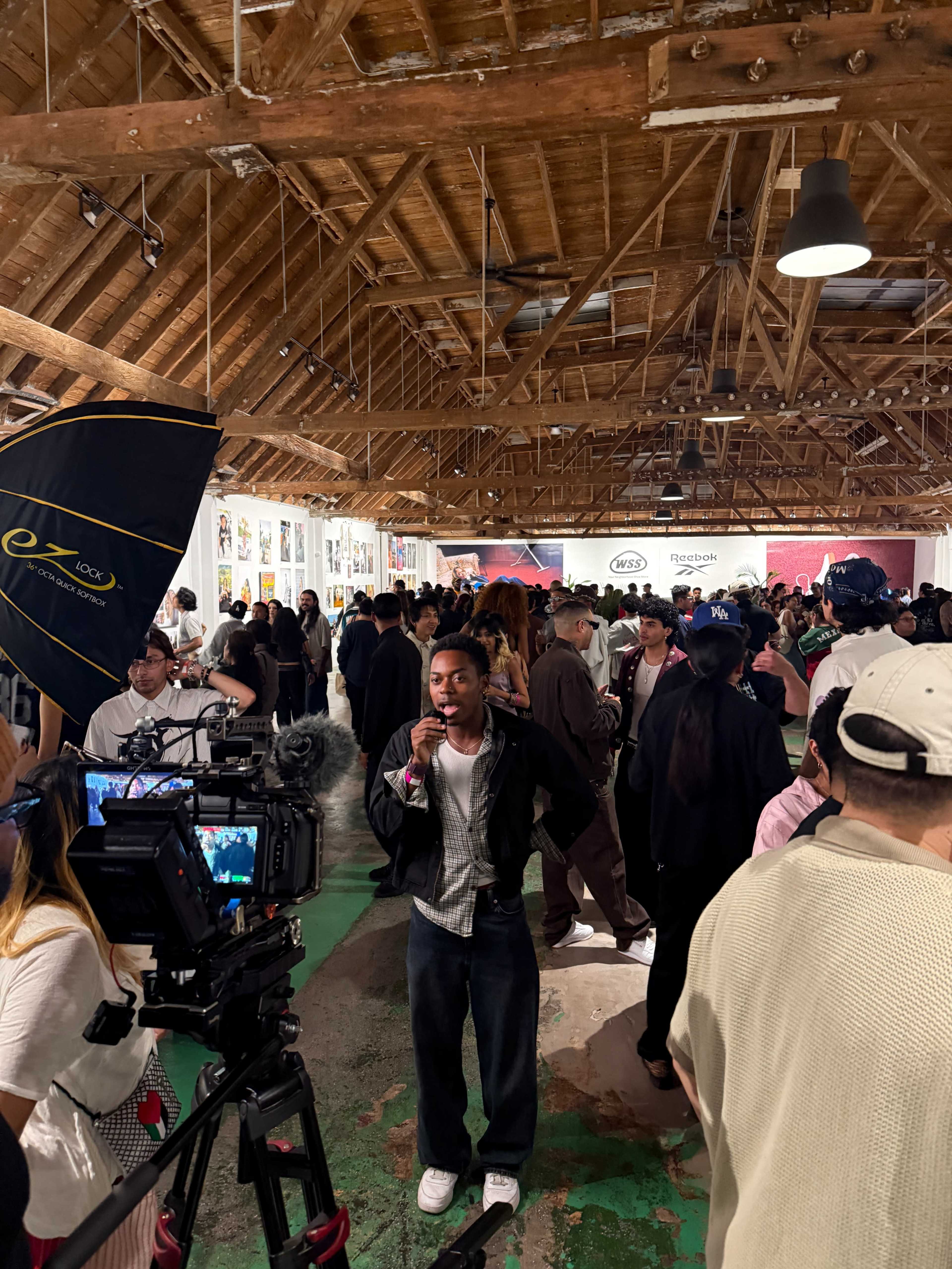 A crowd of people gathers inside a spacious art gallery with a wooden ceiling and multiple artworks displayed on the walls.