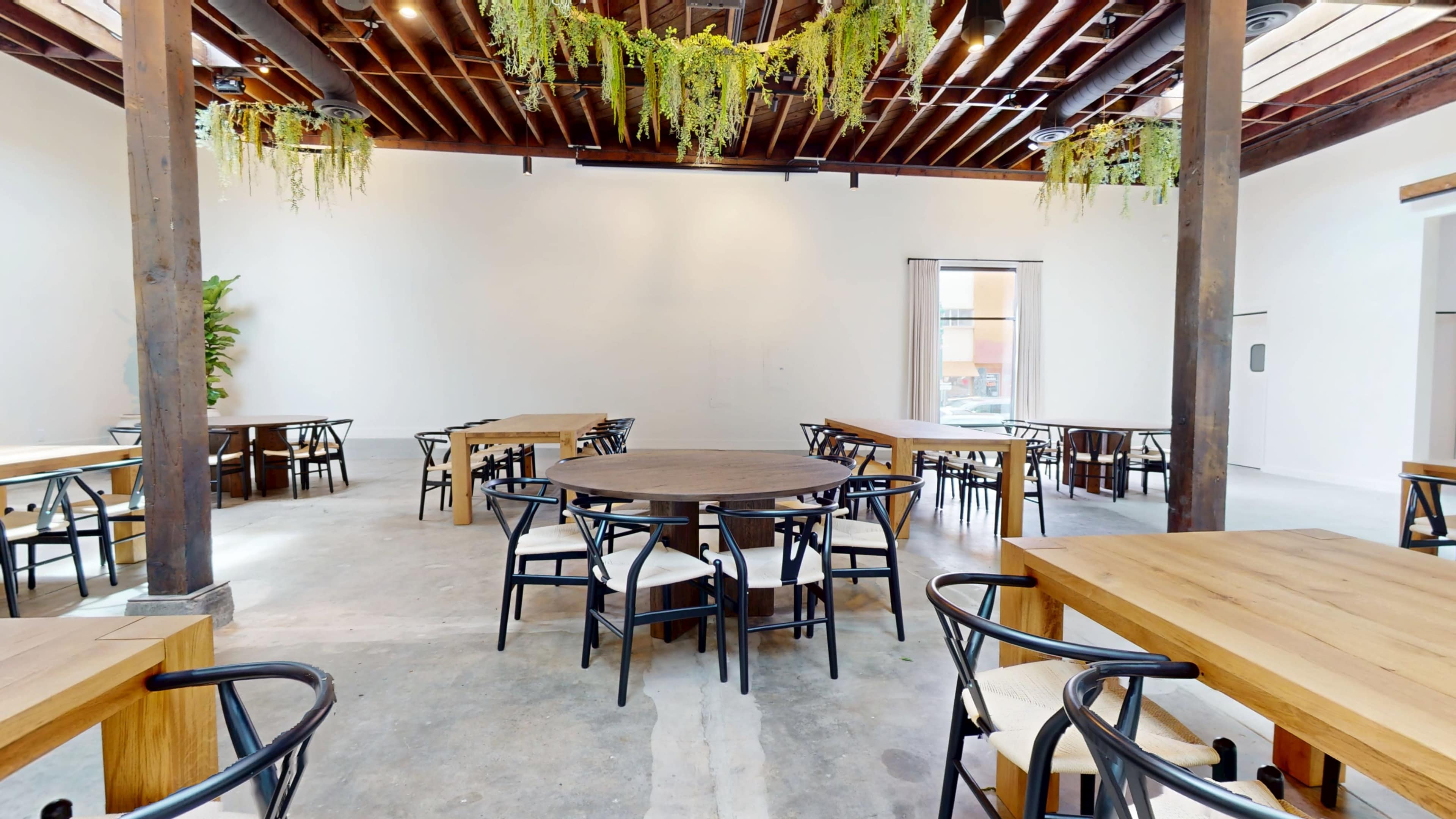 The image shows a spacious dining area with wooden tables and chairs arranged centrally, surrounded by white walls and wooden beams.