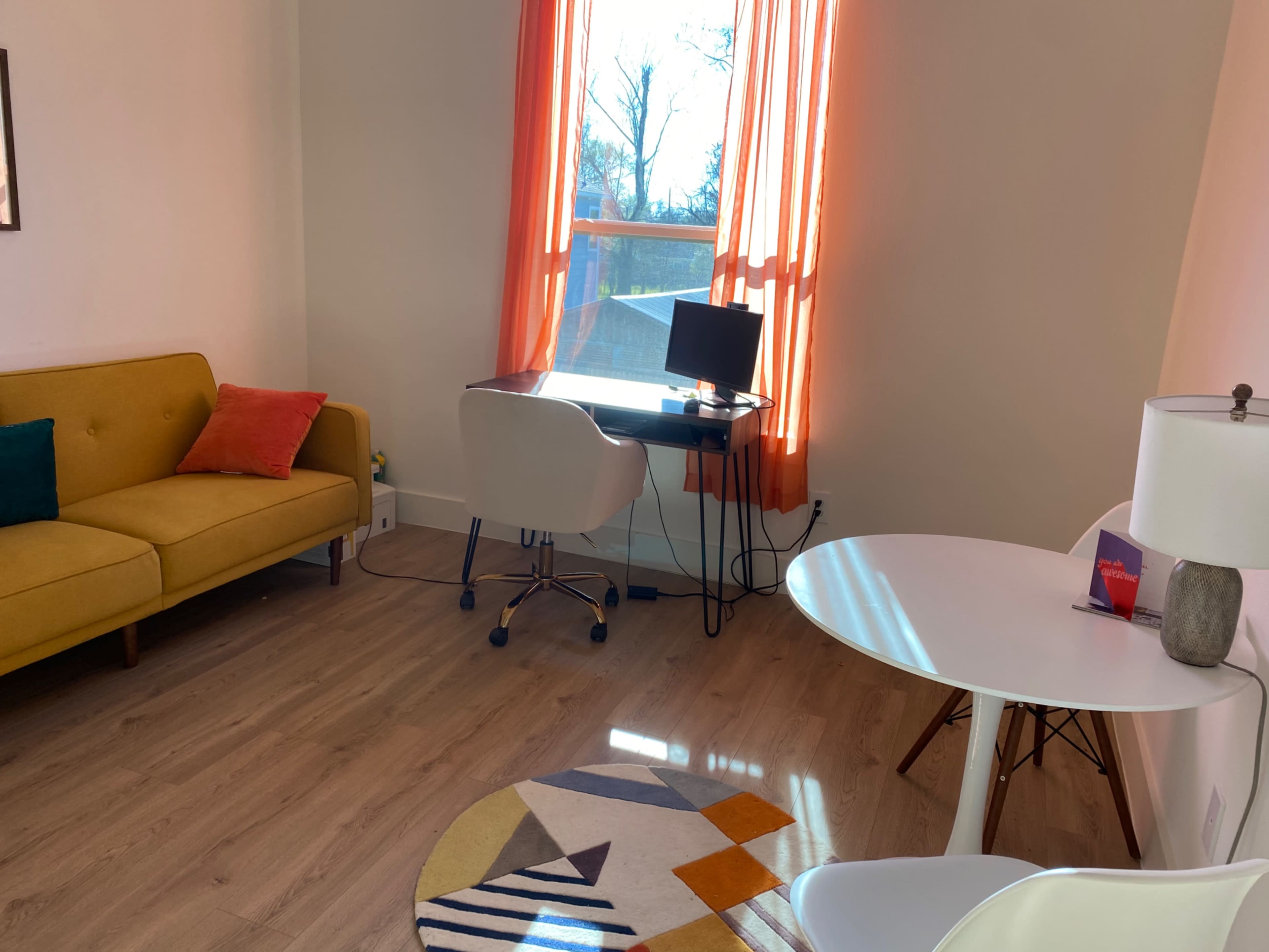 A bright, minimalist living space featuring a yellow sofa, a white desk with a computer, and a round area rug on a light wood floor.