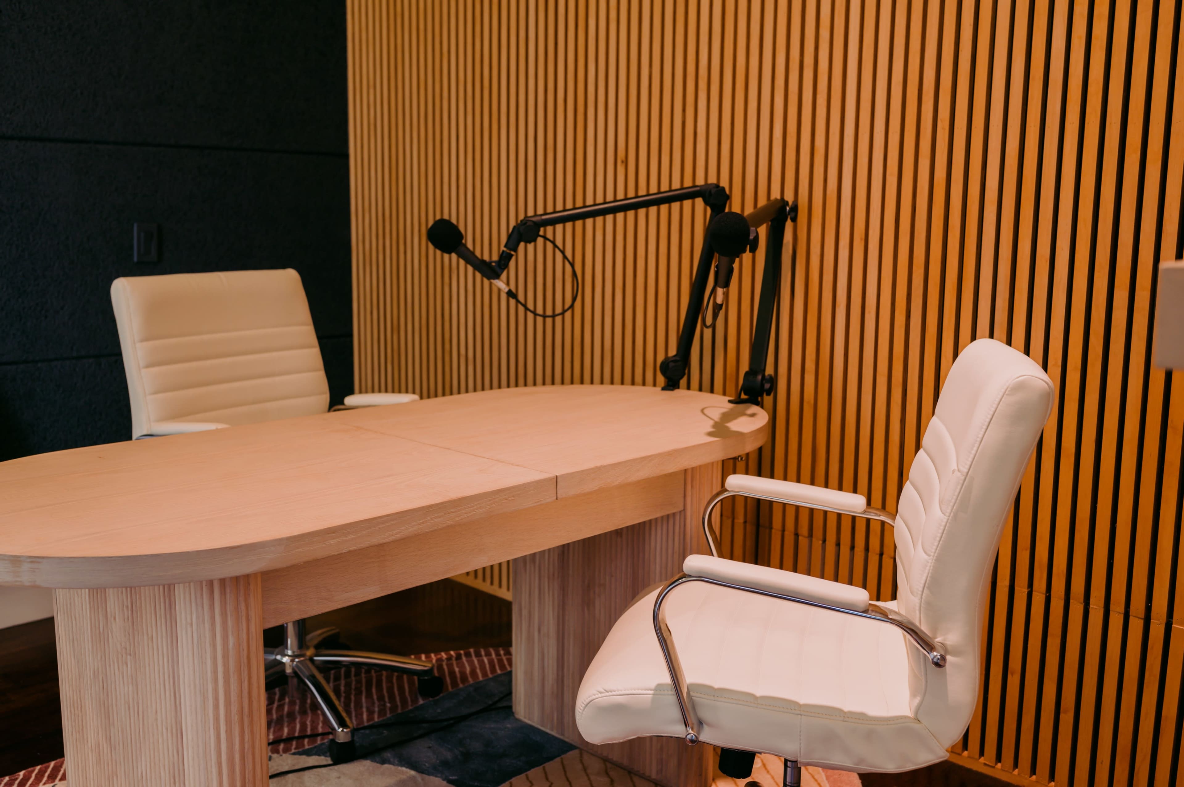 The image shows a modern conference room with a wooden table, two white chairs, and a microphone mounted on an arm against a wooden slat wall.