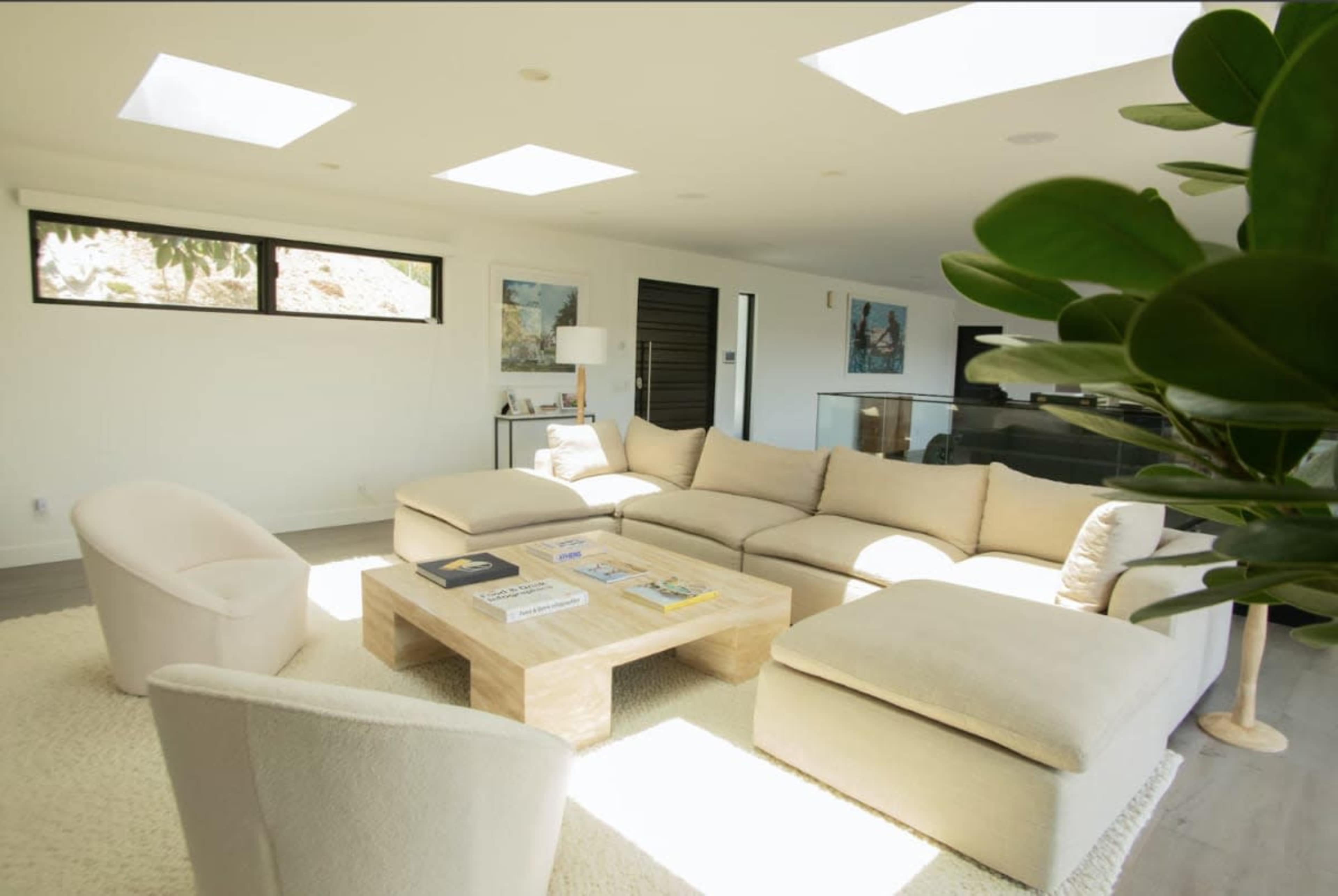 The image depicts a modern living room with a large sectional sofa, a wooden coffee table, and natural light streaming in through skylights.