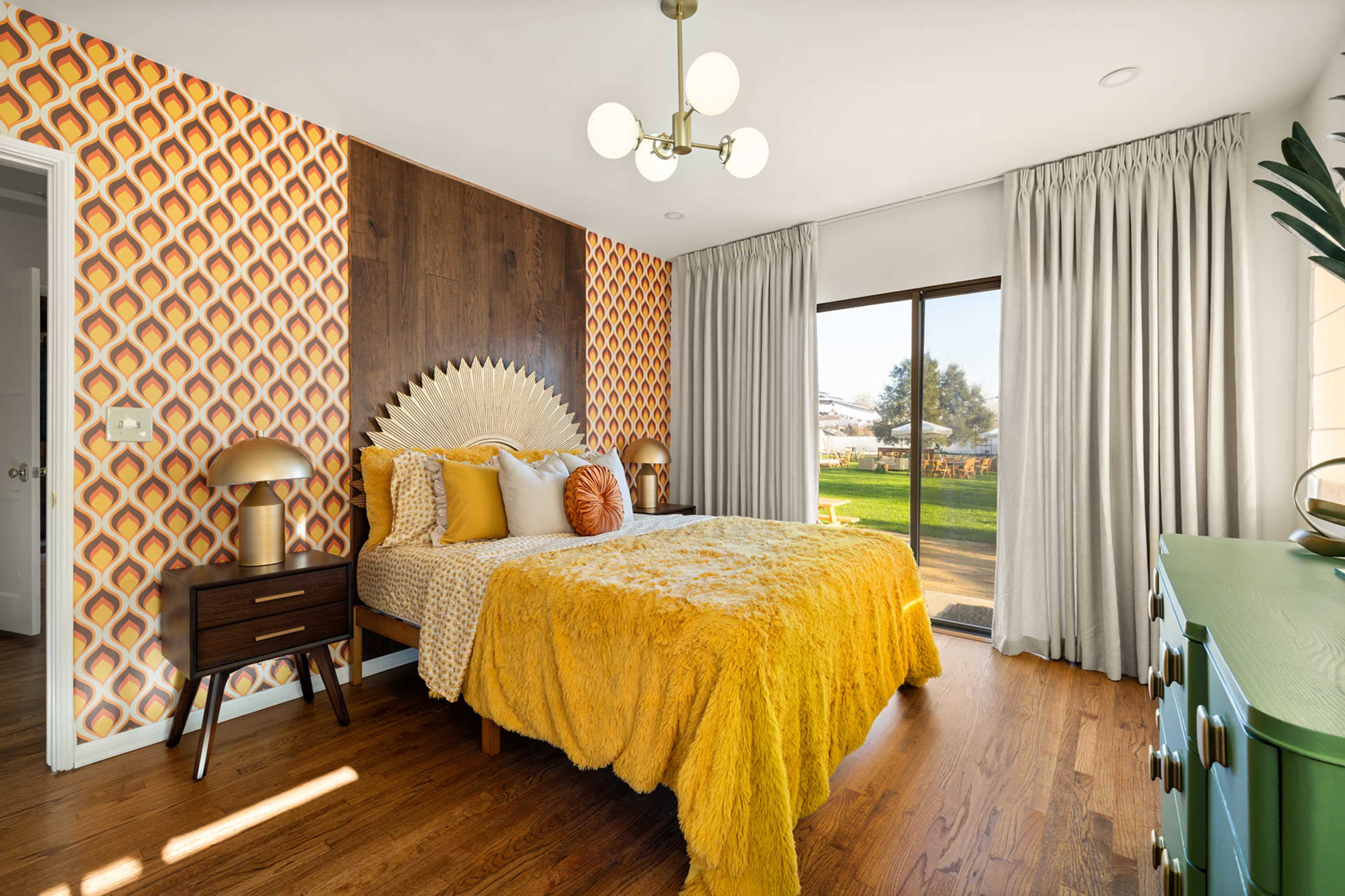 The room features a double bed with a yellow blanket, patterned wallpaper, and large windows overlooking a green outdoor space.