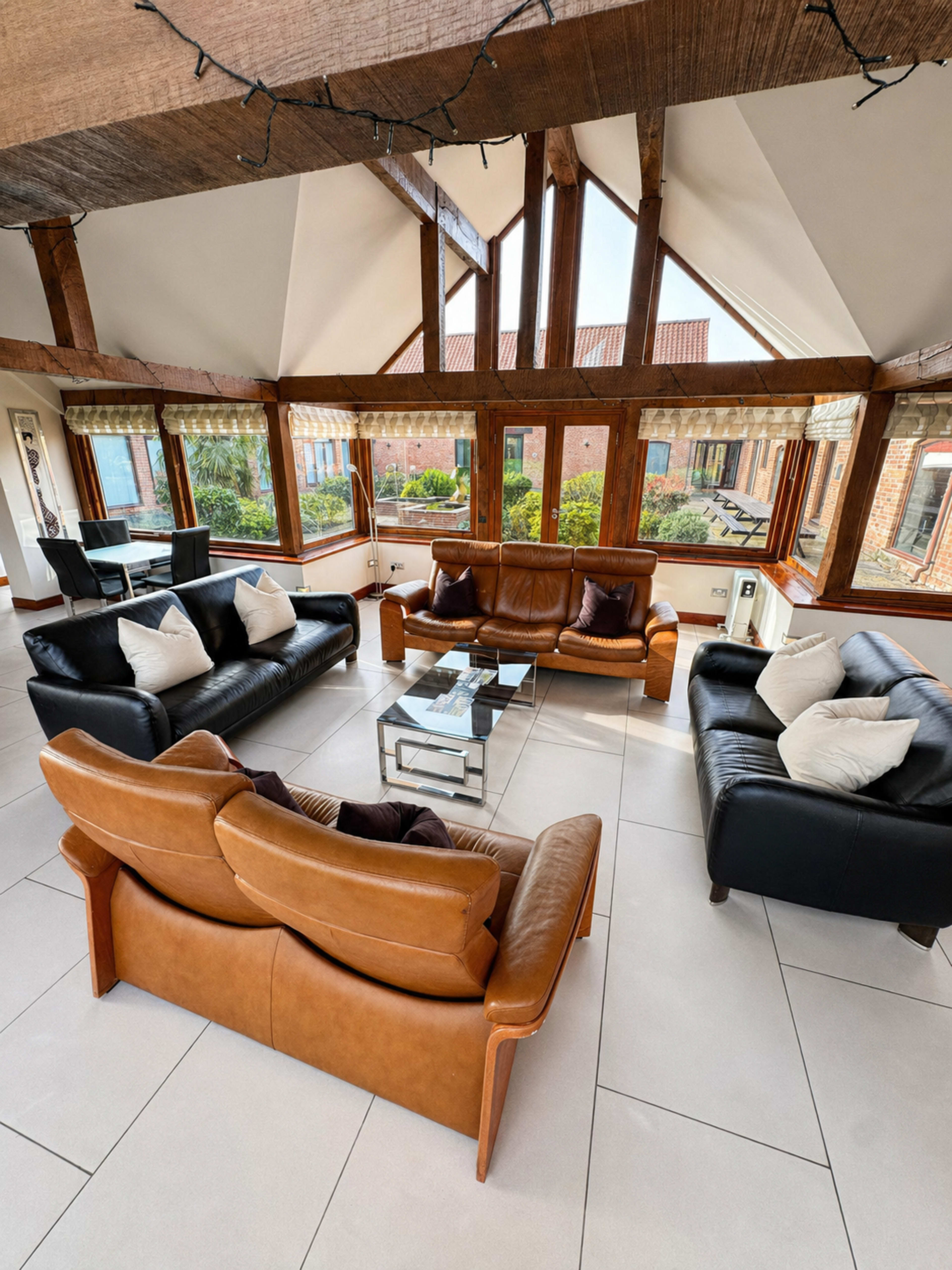 The image shows a spacious living room with large windows, featuring a mix of black and brown leather sofas arranged around a glass coffee table on tiled flooring.