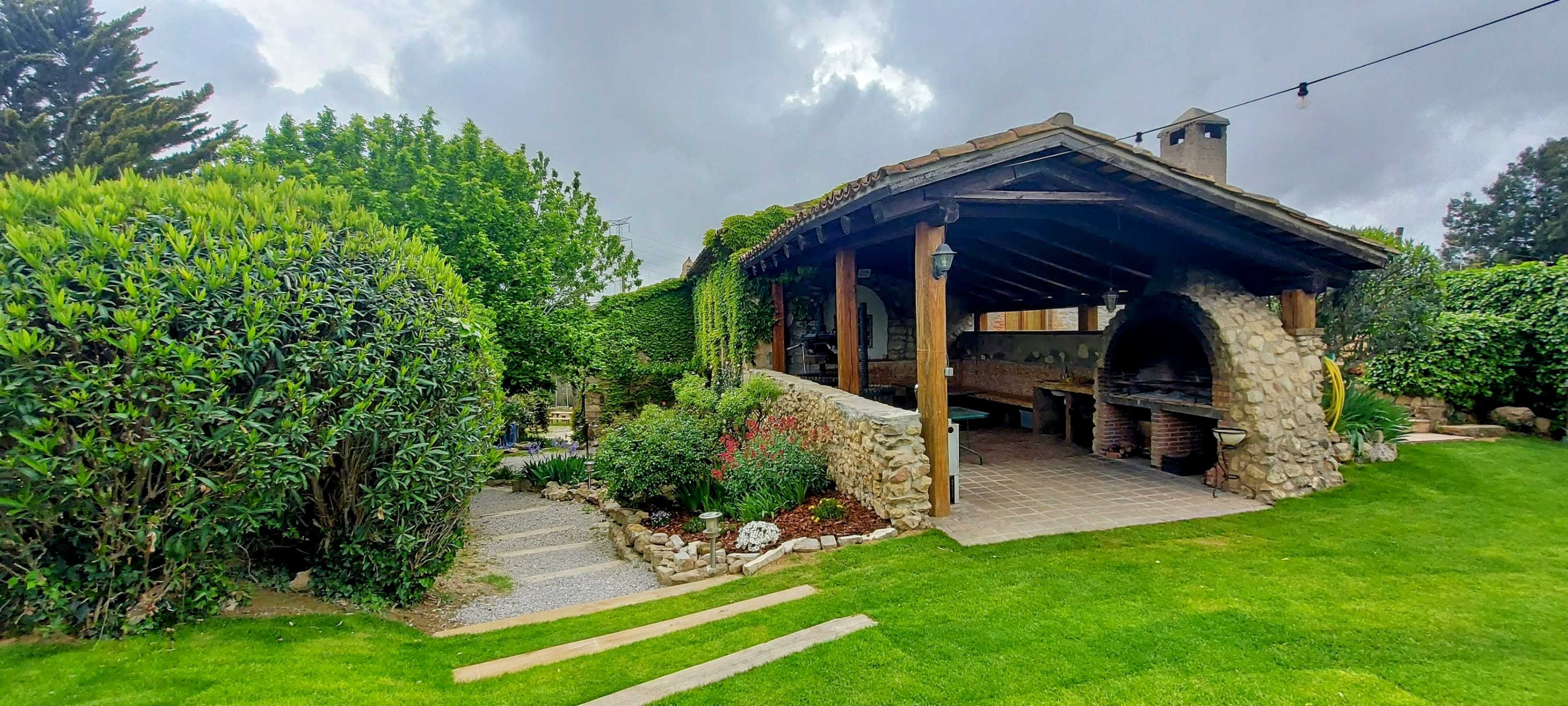 A stone-covered outdoor kitchen is situated under a wooden structure surrounded by lush greenery and a well-maintained garden.