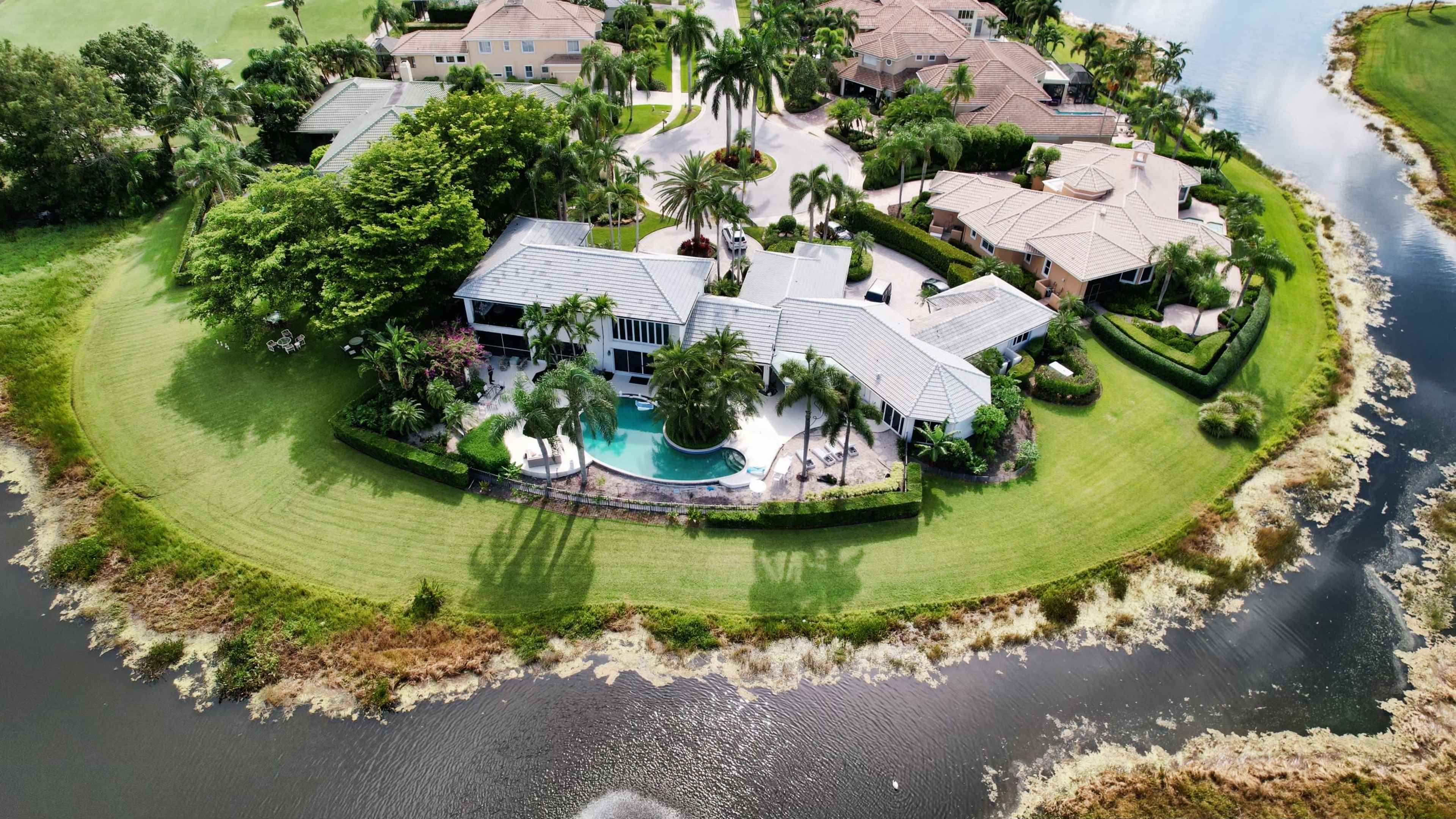 Aerial view of a waterfront property surrounded by a lush lawn and palm trees, with a pool and several nearby houses.