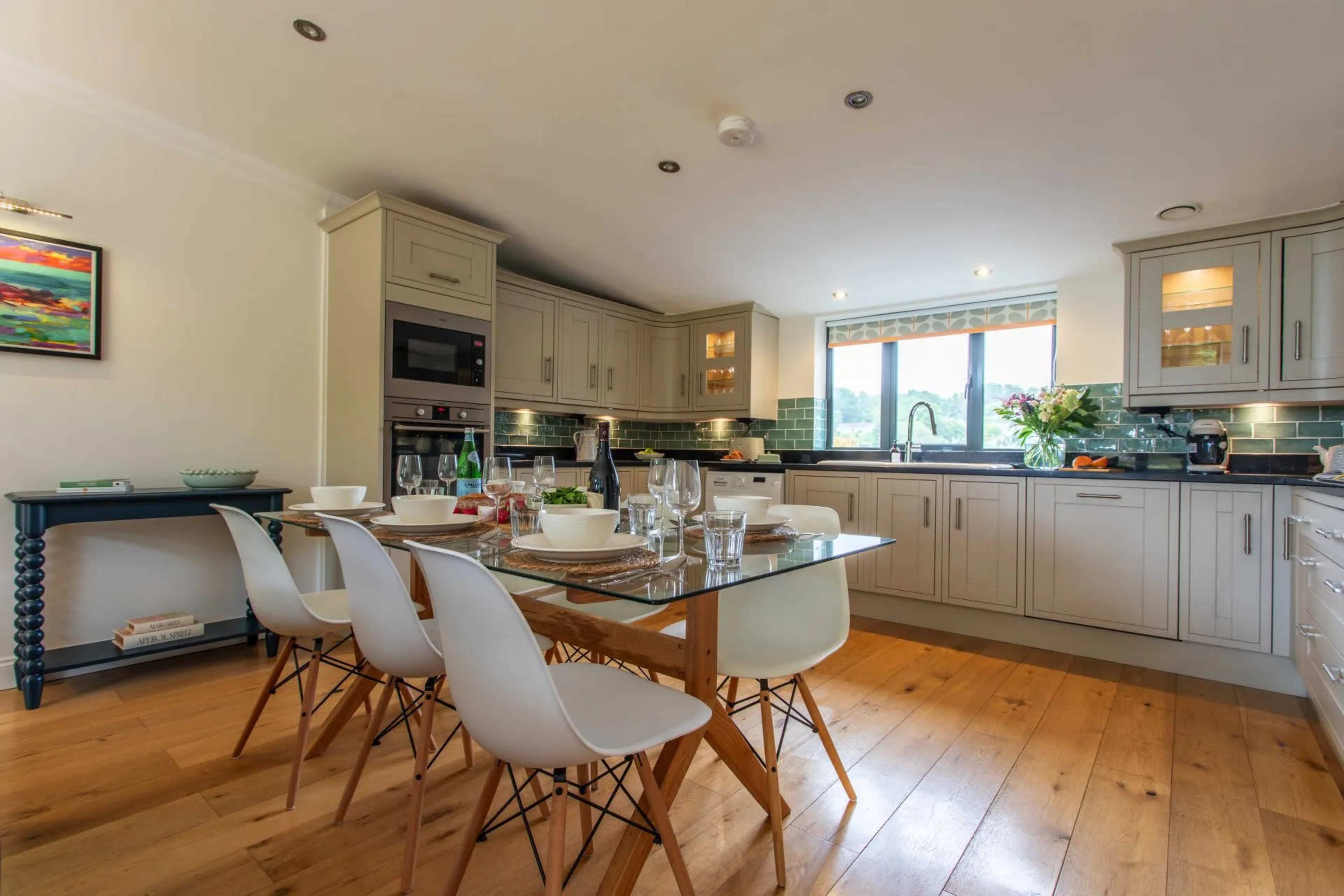 A modern kitchen features a glass dining table with white chairs, surrounded by cabinets and appliances, with natural light coming through a window.