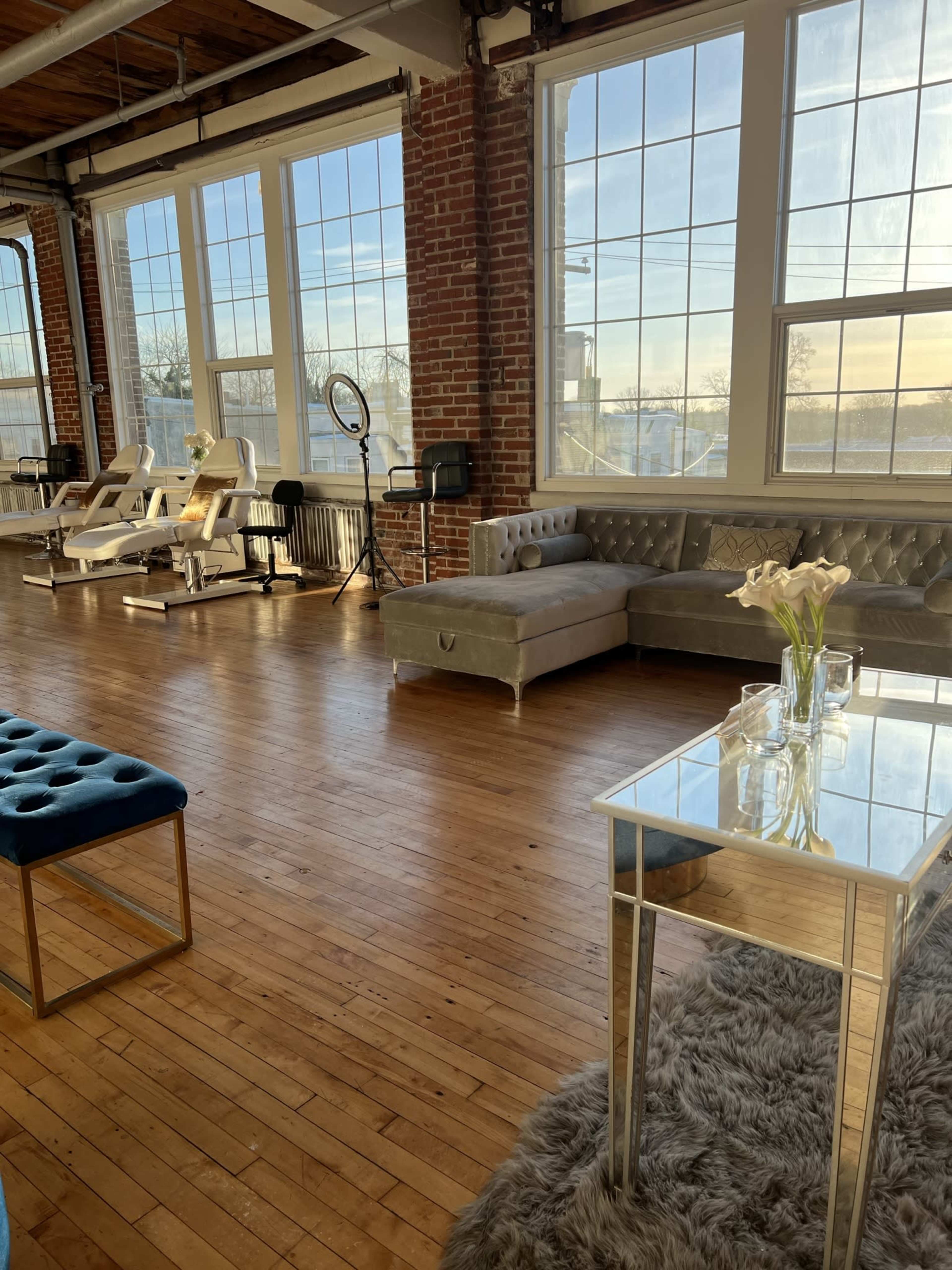 The image shows a bright, open studio space with wooden floors, large windows, a gray couch, a glass table, and various beauty equipment.
