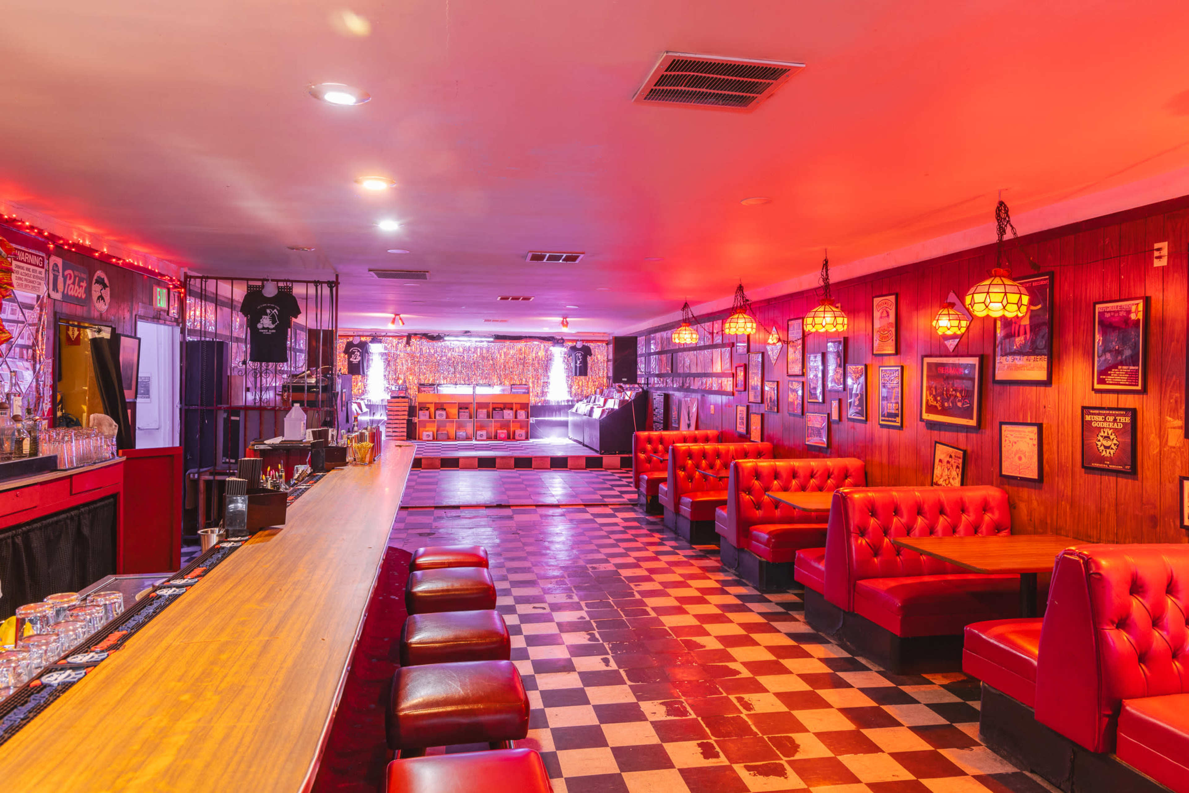 A bar with a long countertop, red padded booths along the sides, and checkered flooring, illuminated by dim red lighting and decorated with framed pictures on the walls.