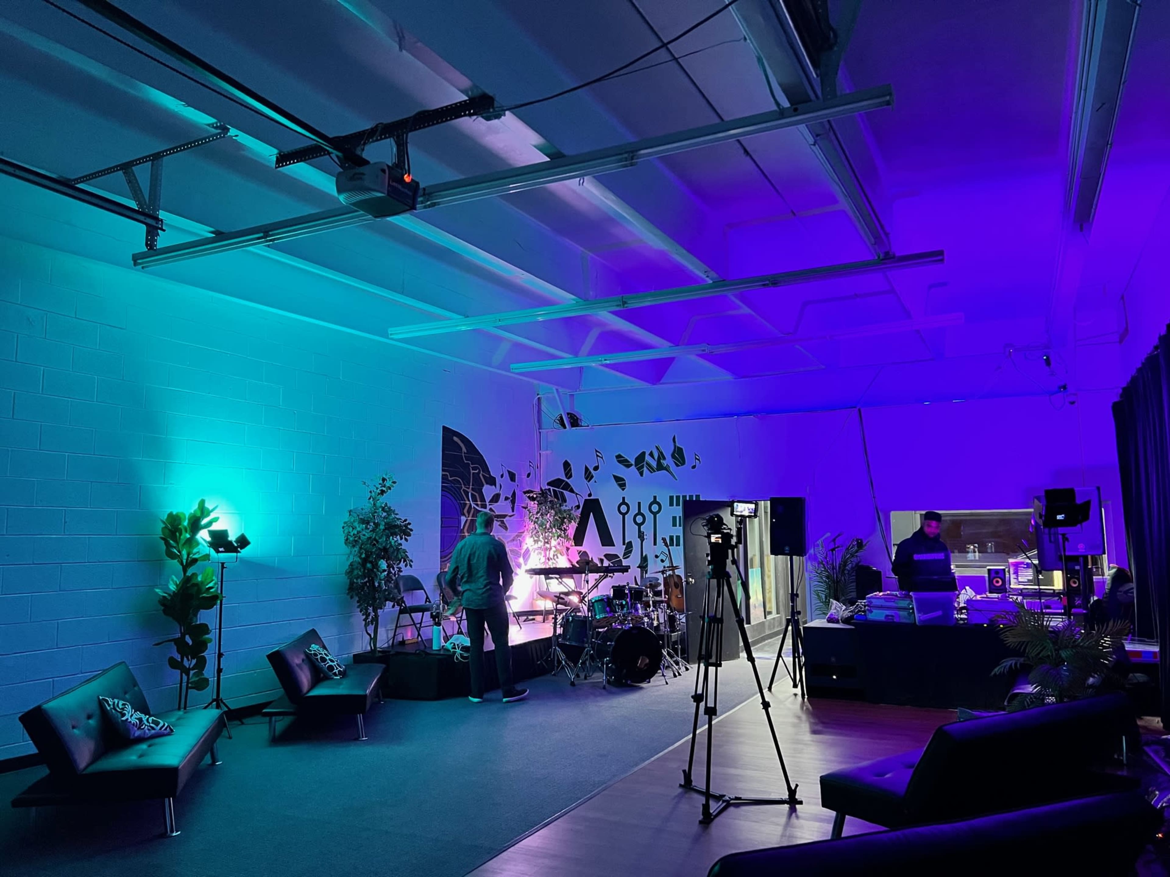 A modern event space with purple and teal lighting, featuring a drum set, couches, and a DJ setup.