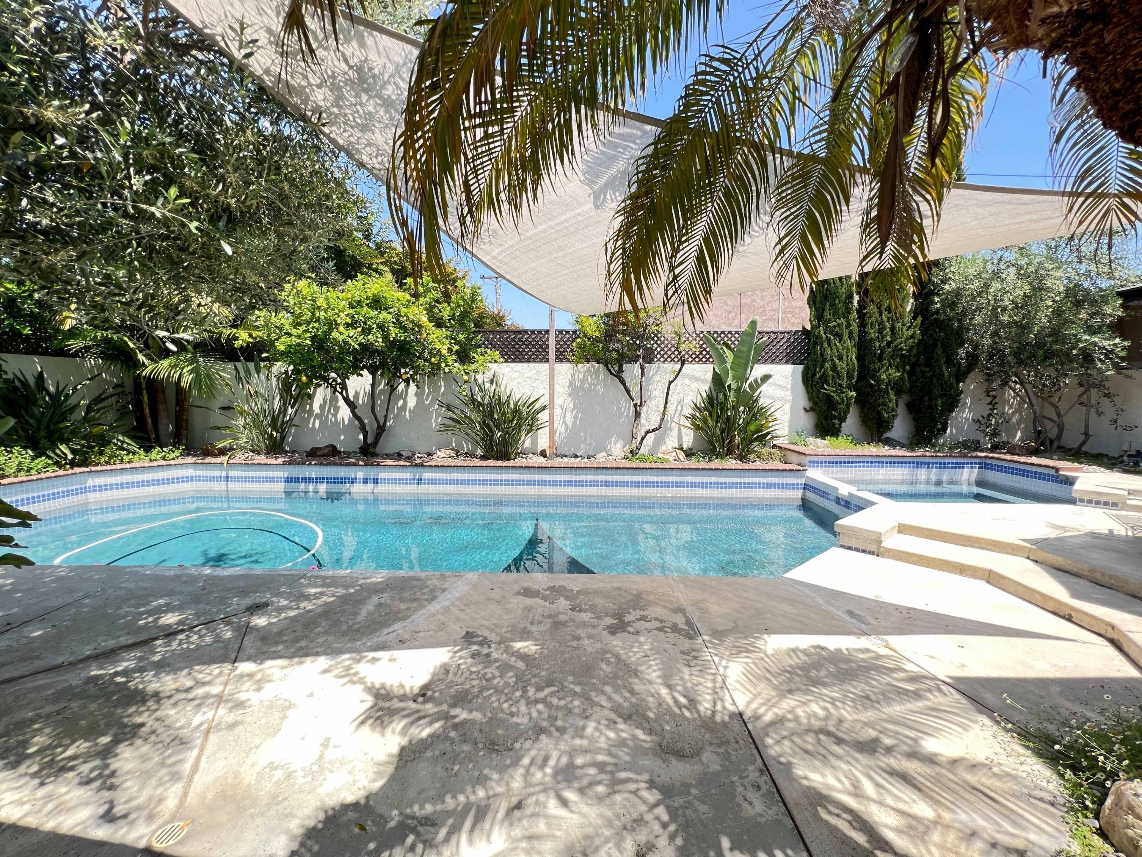 A swimming pool is surrounded by greenery, with shaded areas provided by fabric canopies overhead.