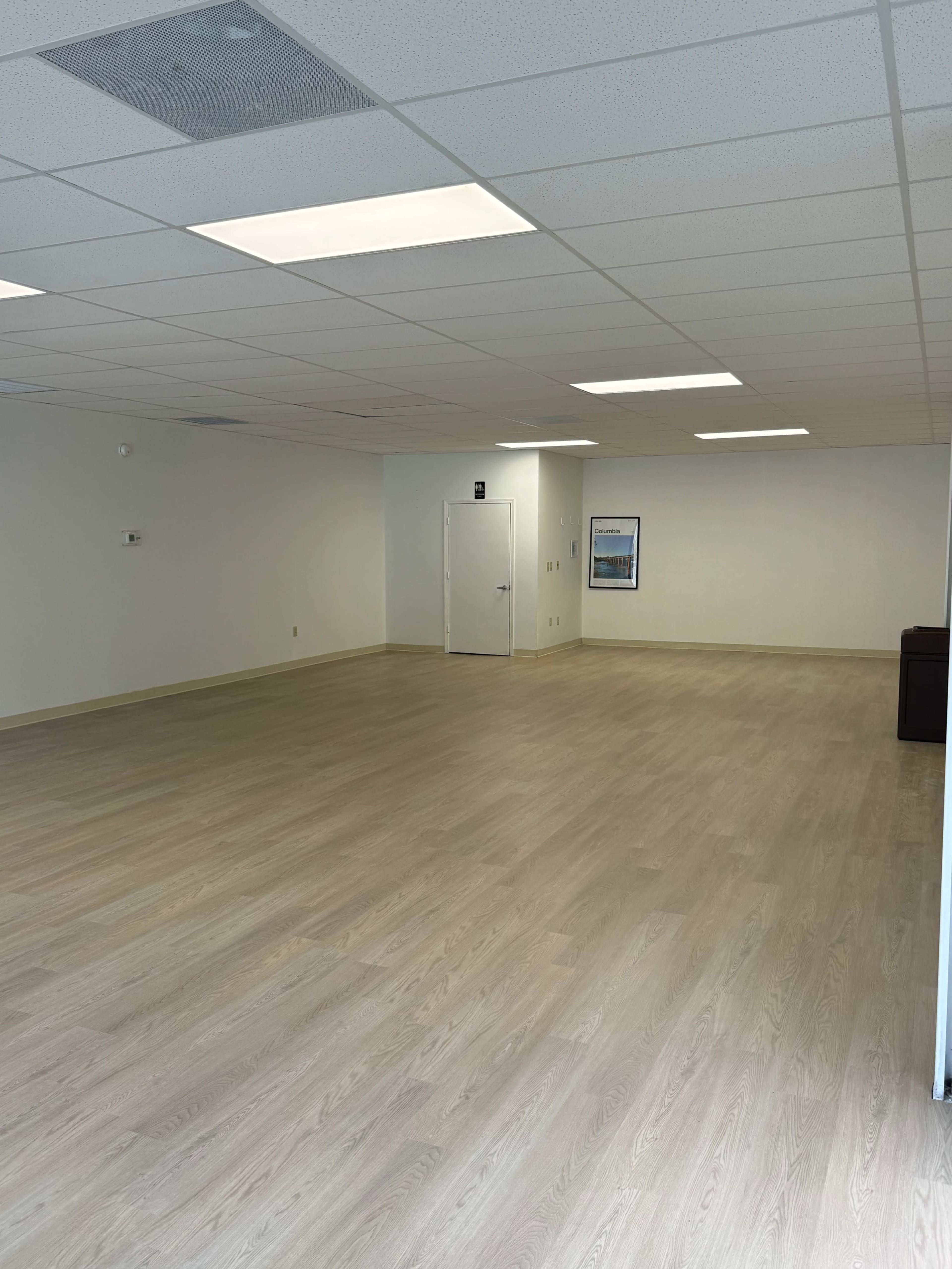 The image shows a spacious, empty room with light-colored wooden flooring and plain white walls, featuring a door on one side and ceiling lights above.