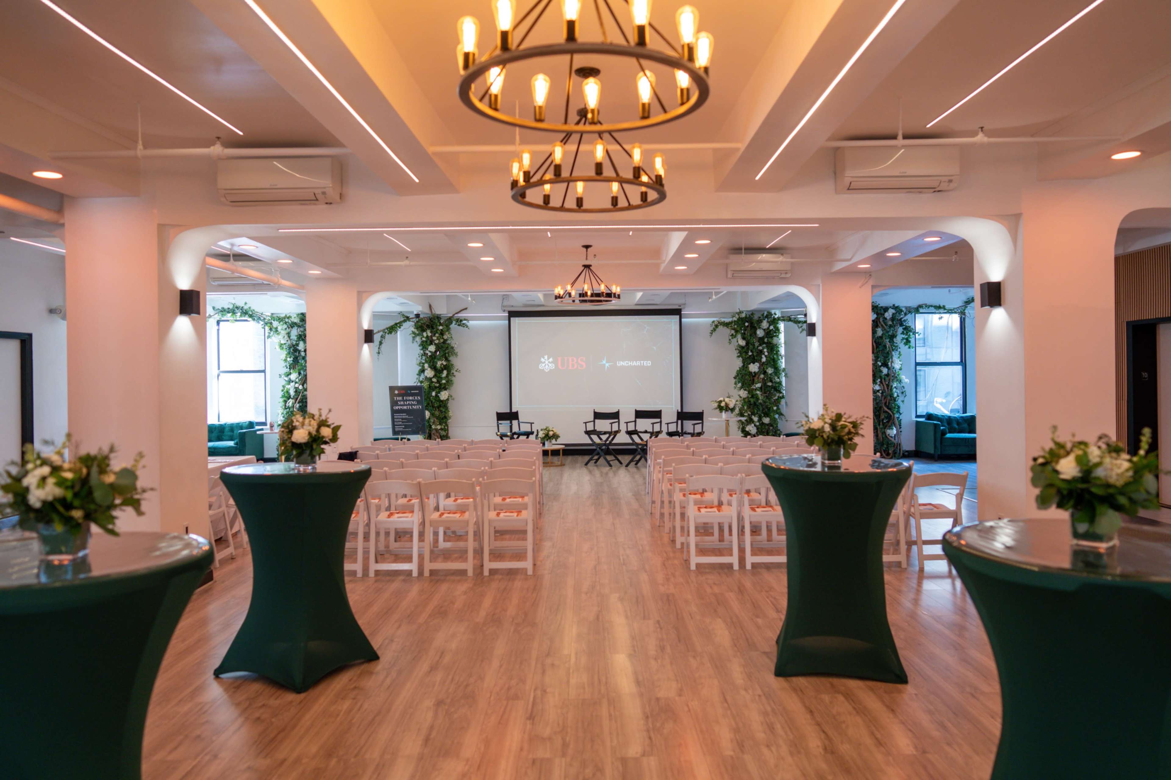 The image shows a spacious event room set up for a gathering, featuring chairs arranged in rows, high cocktail tables, and greenery decorating the space.
