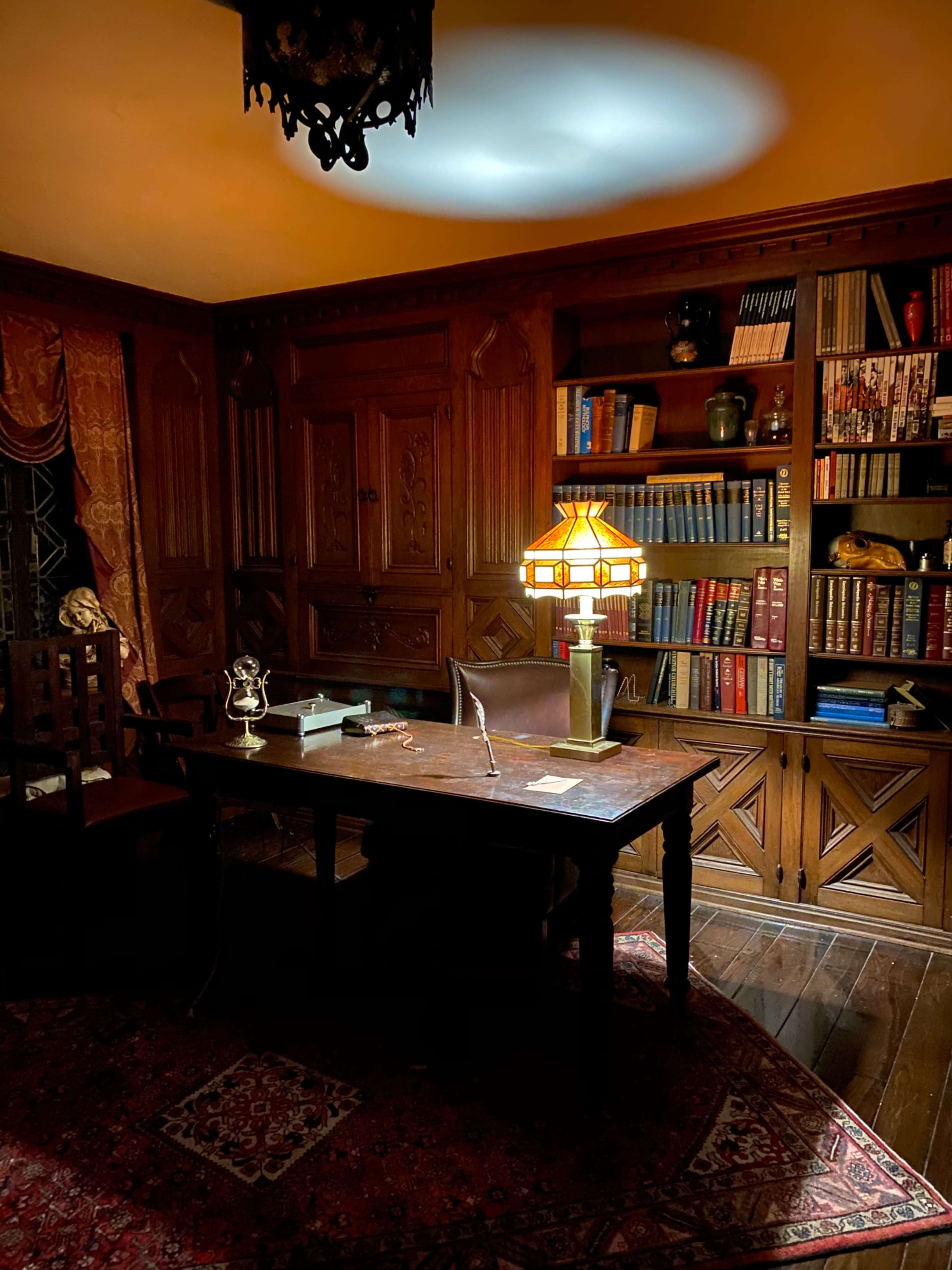 A dimly lit wooden office with a large desk, a classic lamp, and a bookshelf filled with various books.