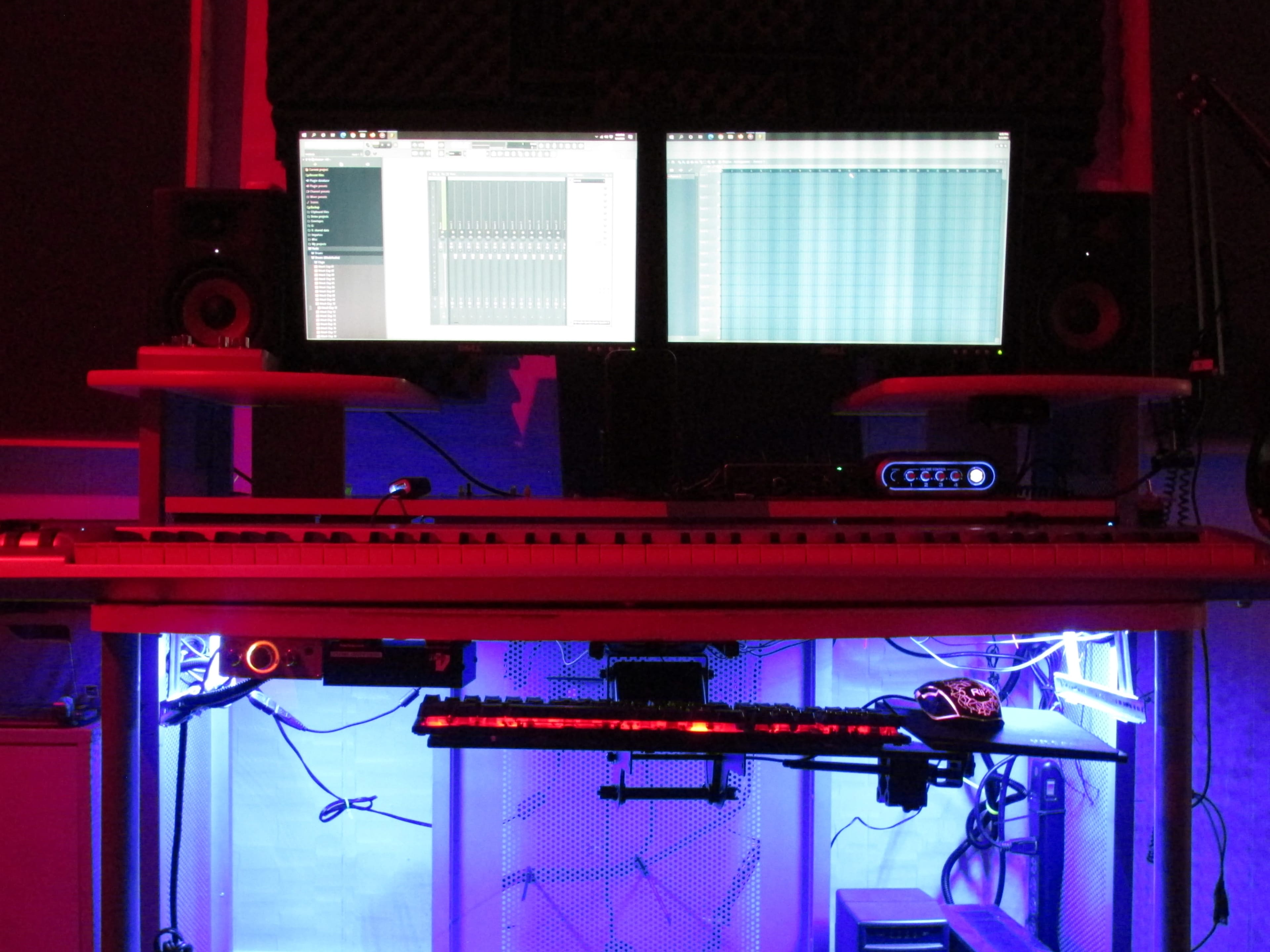 A music production setup features two monitors displaying software, a keyboard, and equipment illuminated by blue and red lighting.