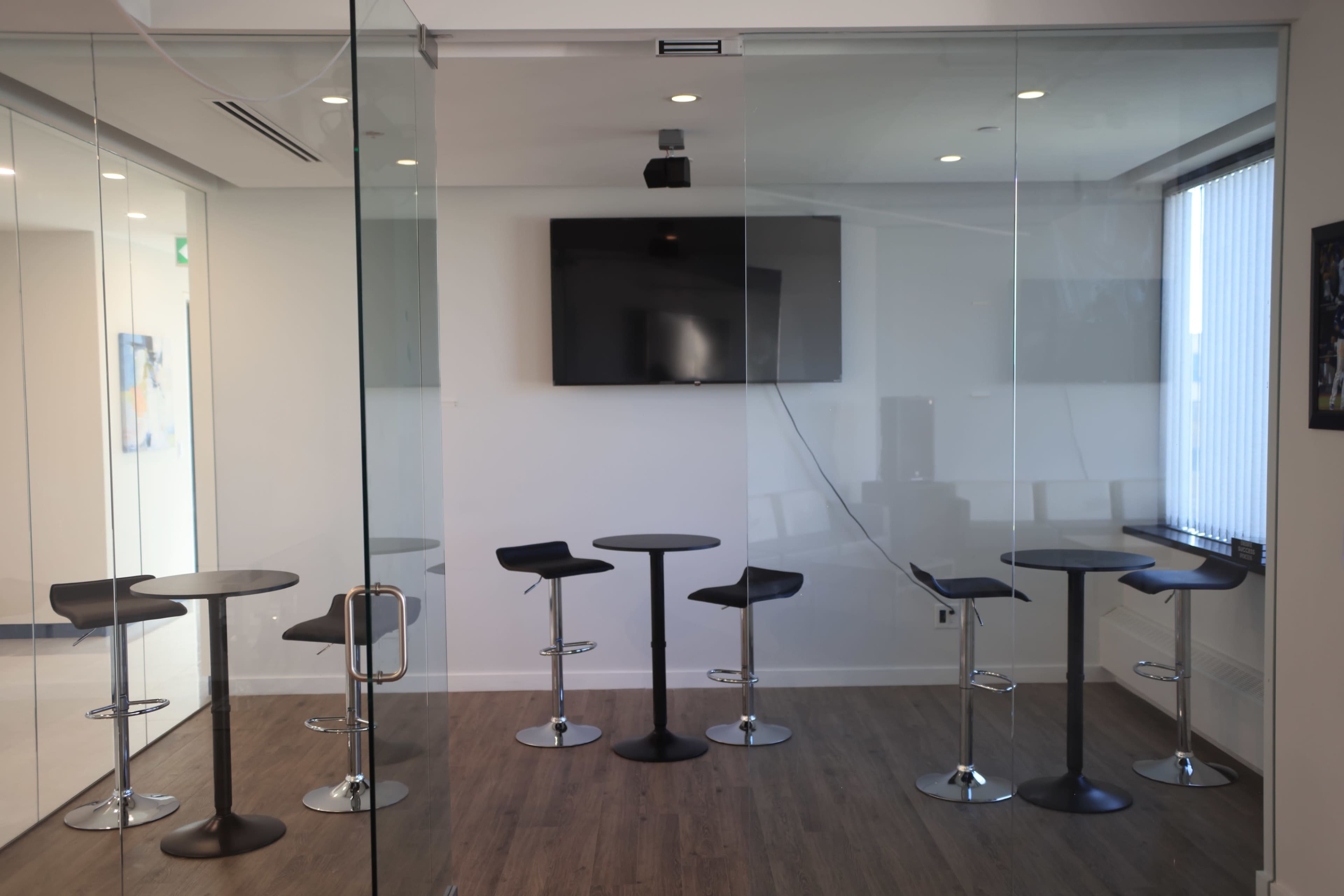 The image shows a modern conference room with glass walls, featuring several tall black and silver bar stools arranged around round tables and a wall-mounted television.