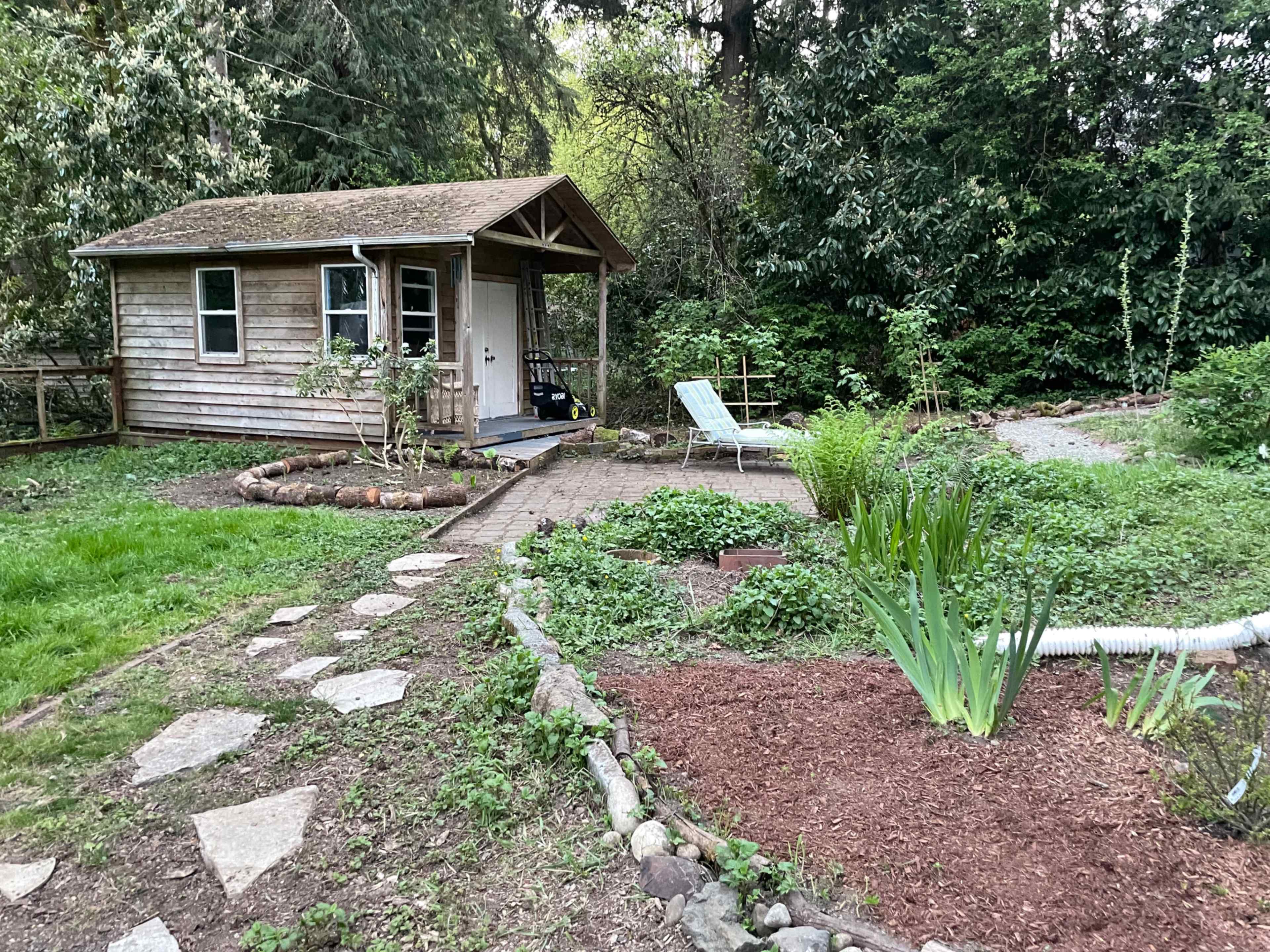 A small wooden cabin sits surrounded by a garden with various plants and a stone pathway leading to it.