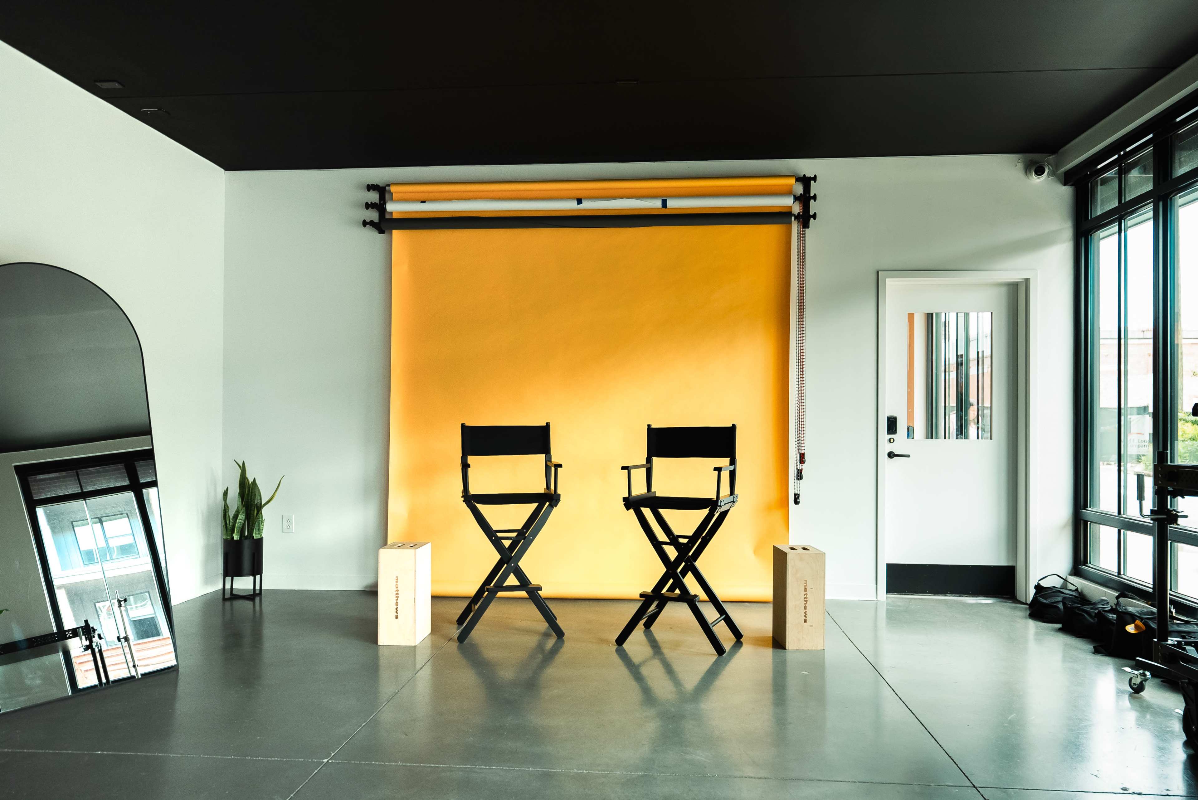 A bright orange backdrop is set up between two director's chairs in a modern studio space with reflective surfaces and a doorway.