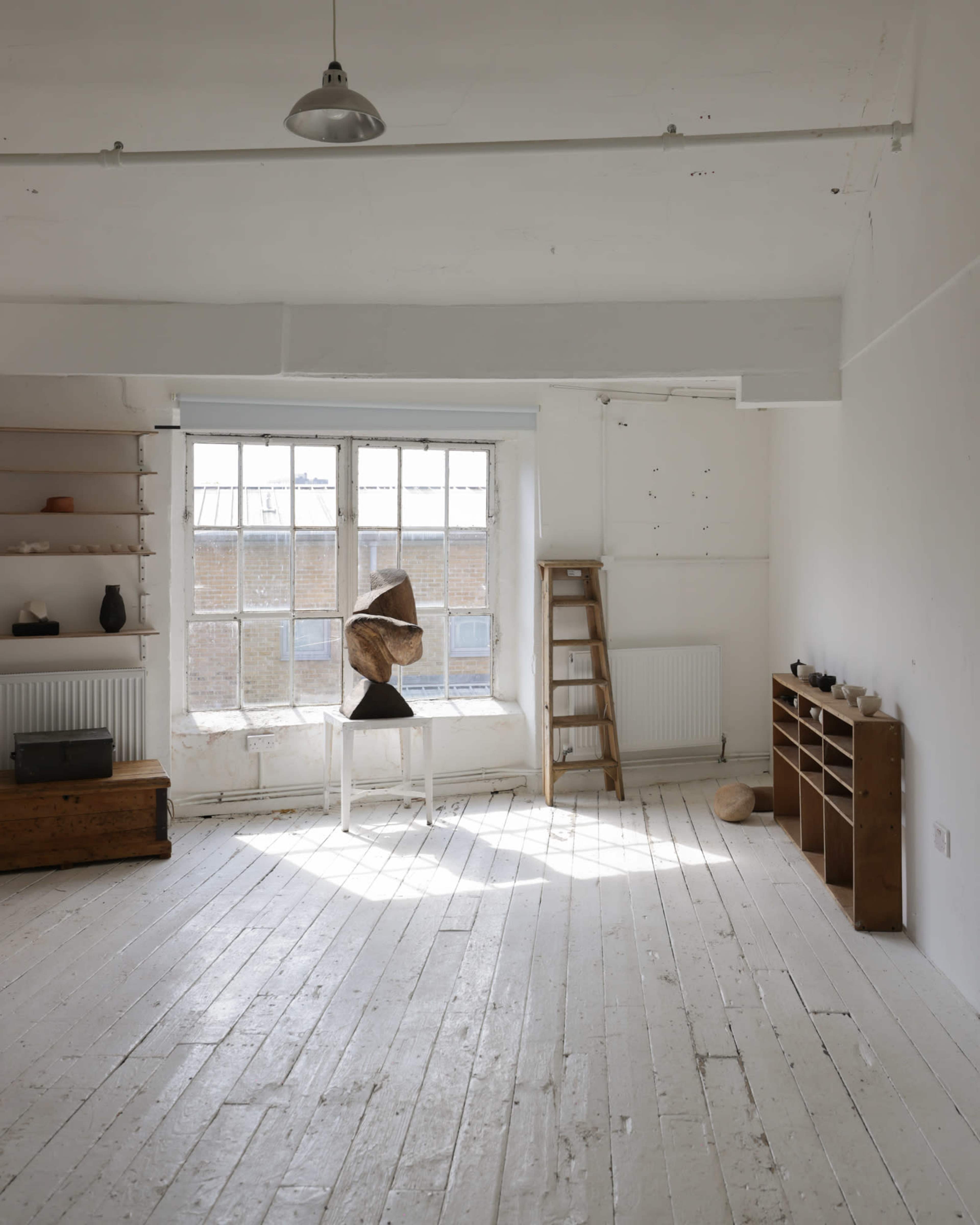 The image shows a spacious, well-lit room with wooden flooring, large windows, and a few pieces of furniture including a sculpture on a white pedestal, a wooden shelf, and a ladder.