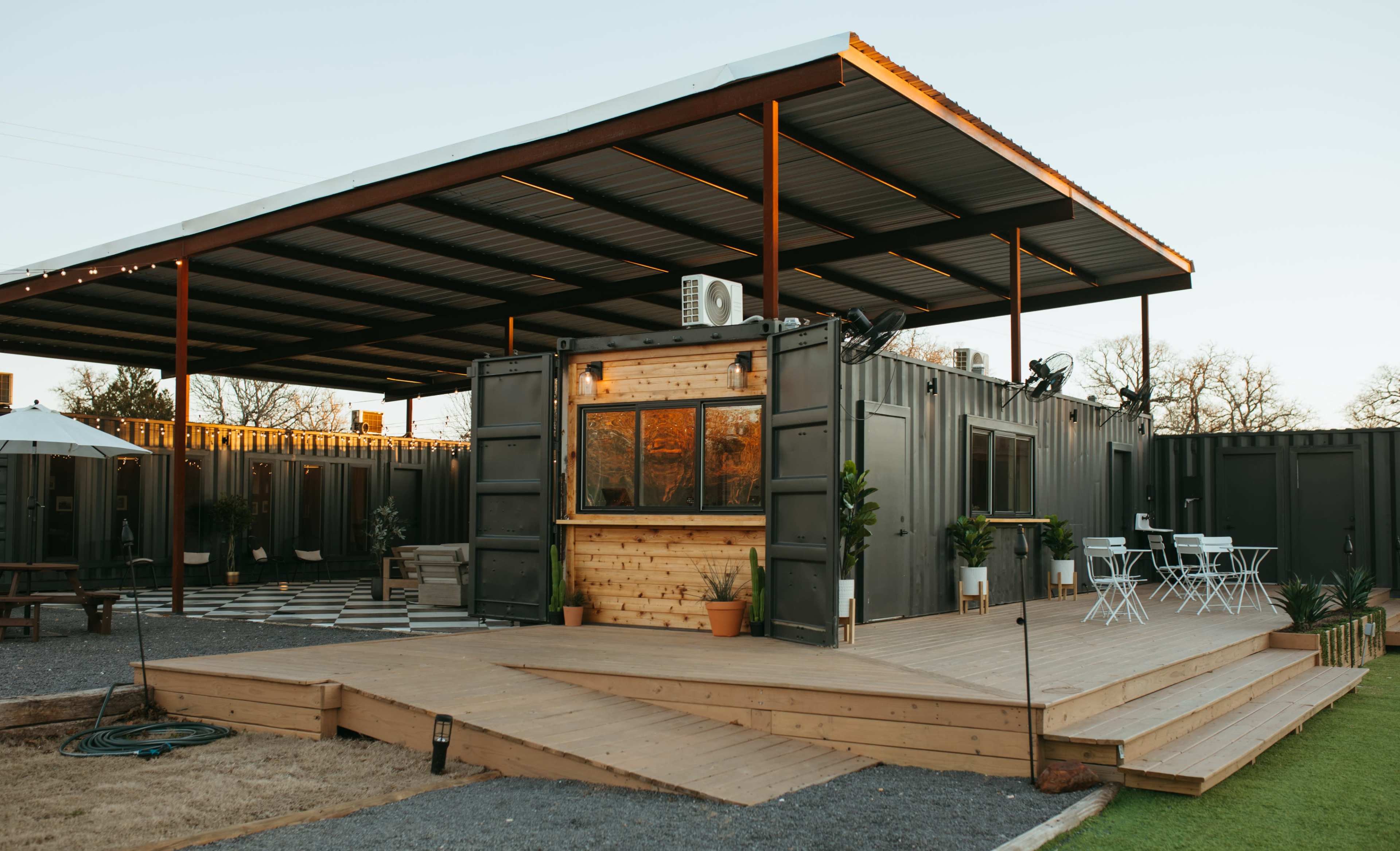 A modern, open-air structure featuring a wooden deck and a container-style building with large windows and outdoor seating.