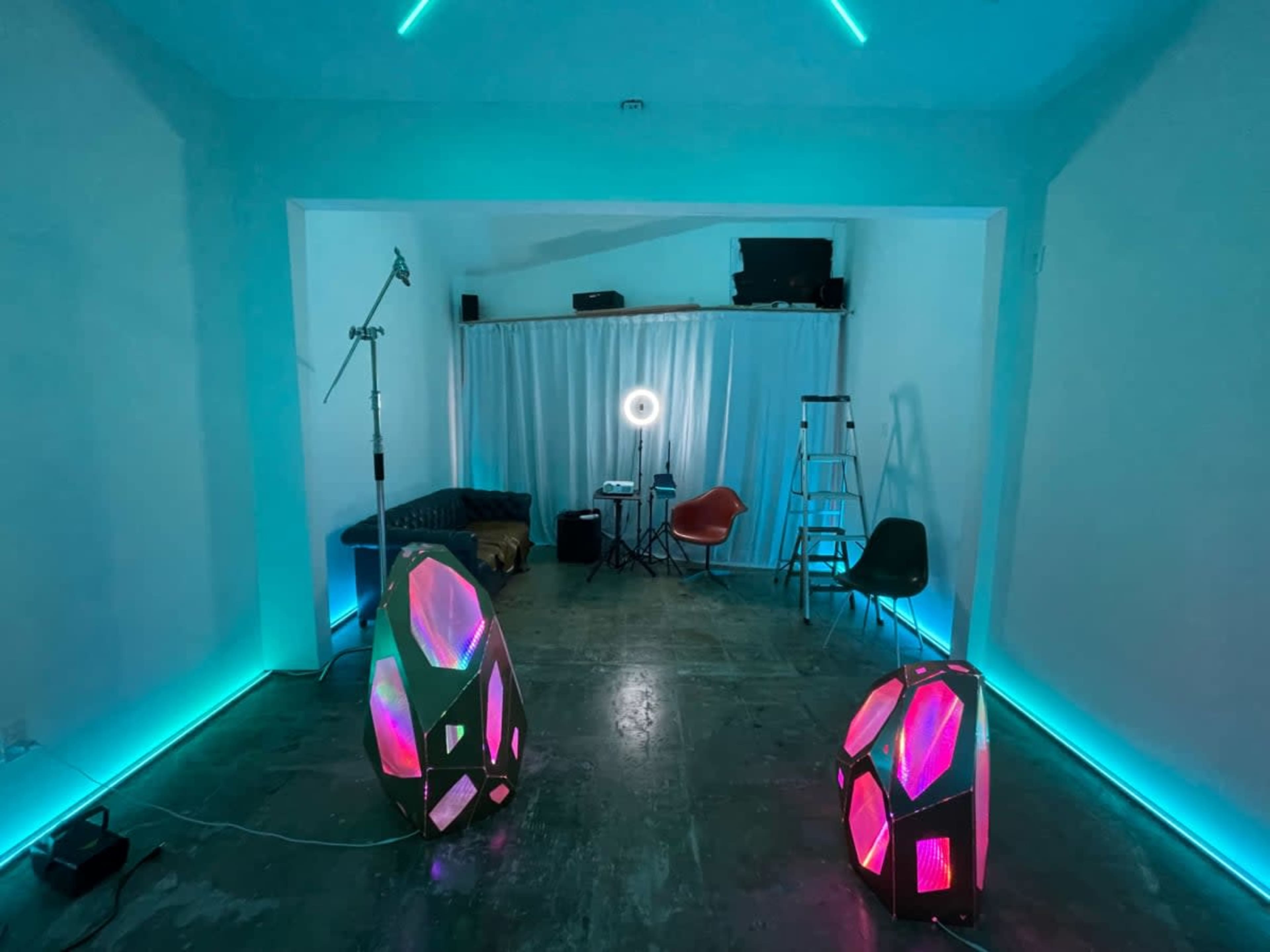 The image shows a minimalistic room featuring two illuminated geometric sculpture-like objects, a couch, a ring light, and a ladder against a backdrop of blue and pink lighting.