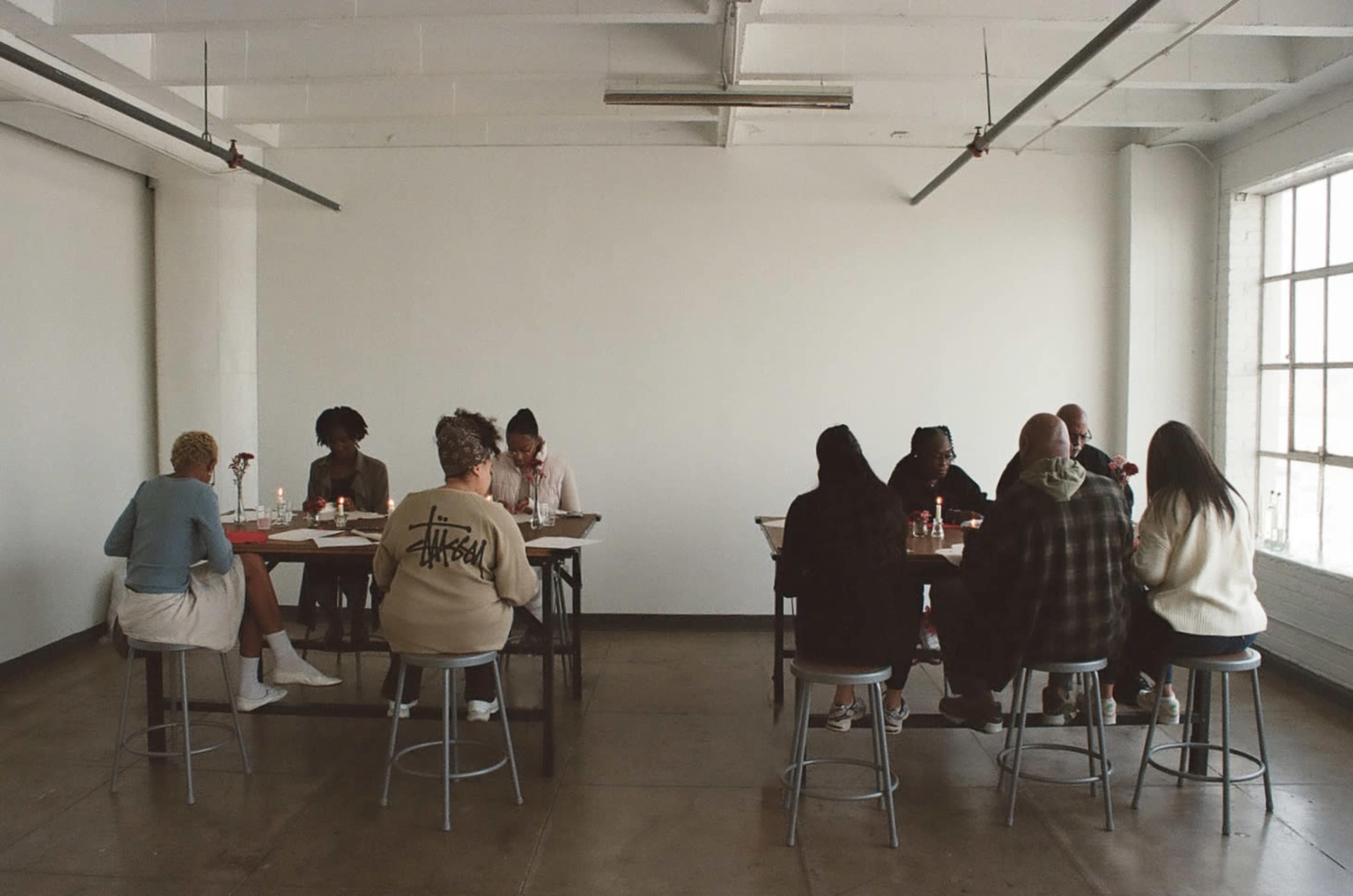 A group of people is seated around several tables in a spacious, bright room, engaged in conversation.