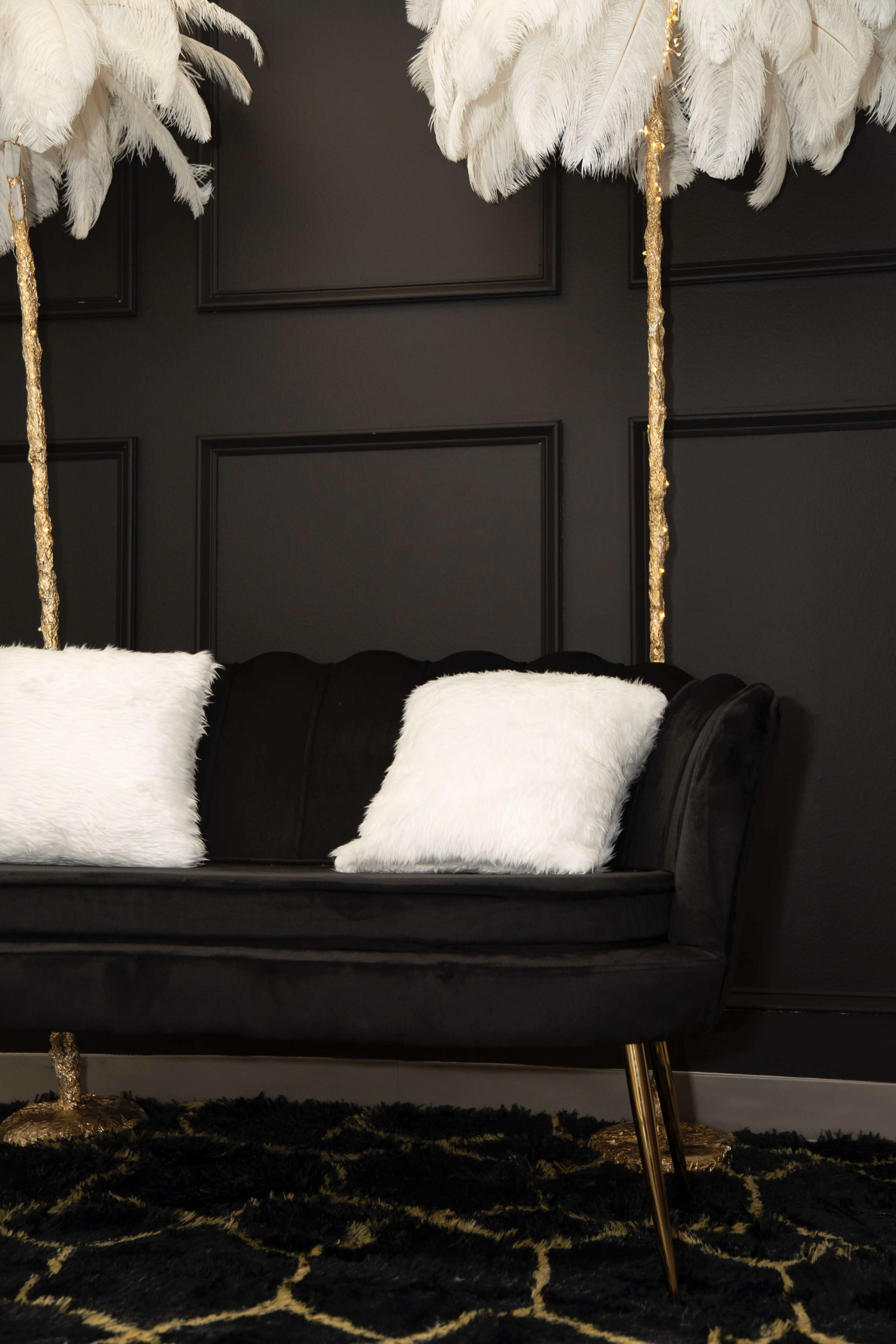 The image shows a black velvet sofa with white pillows positioned between two tall feathered floor lamps against a dark wall.