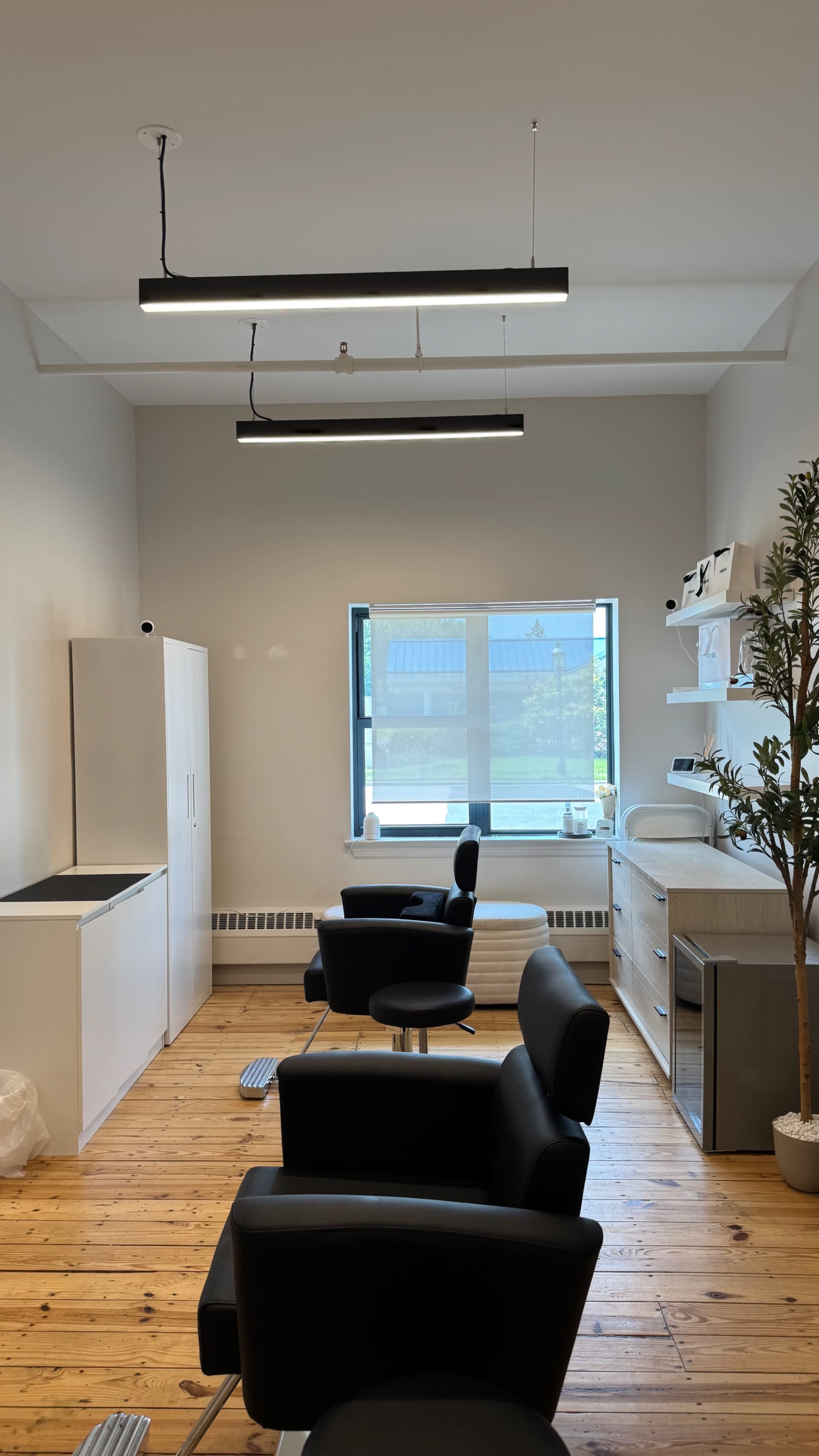 The image shows a well-lit salon interior featuring two black styling chairs, a white cabinetry with a sink, and a large window allowing natural light to enter.