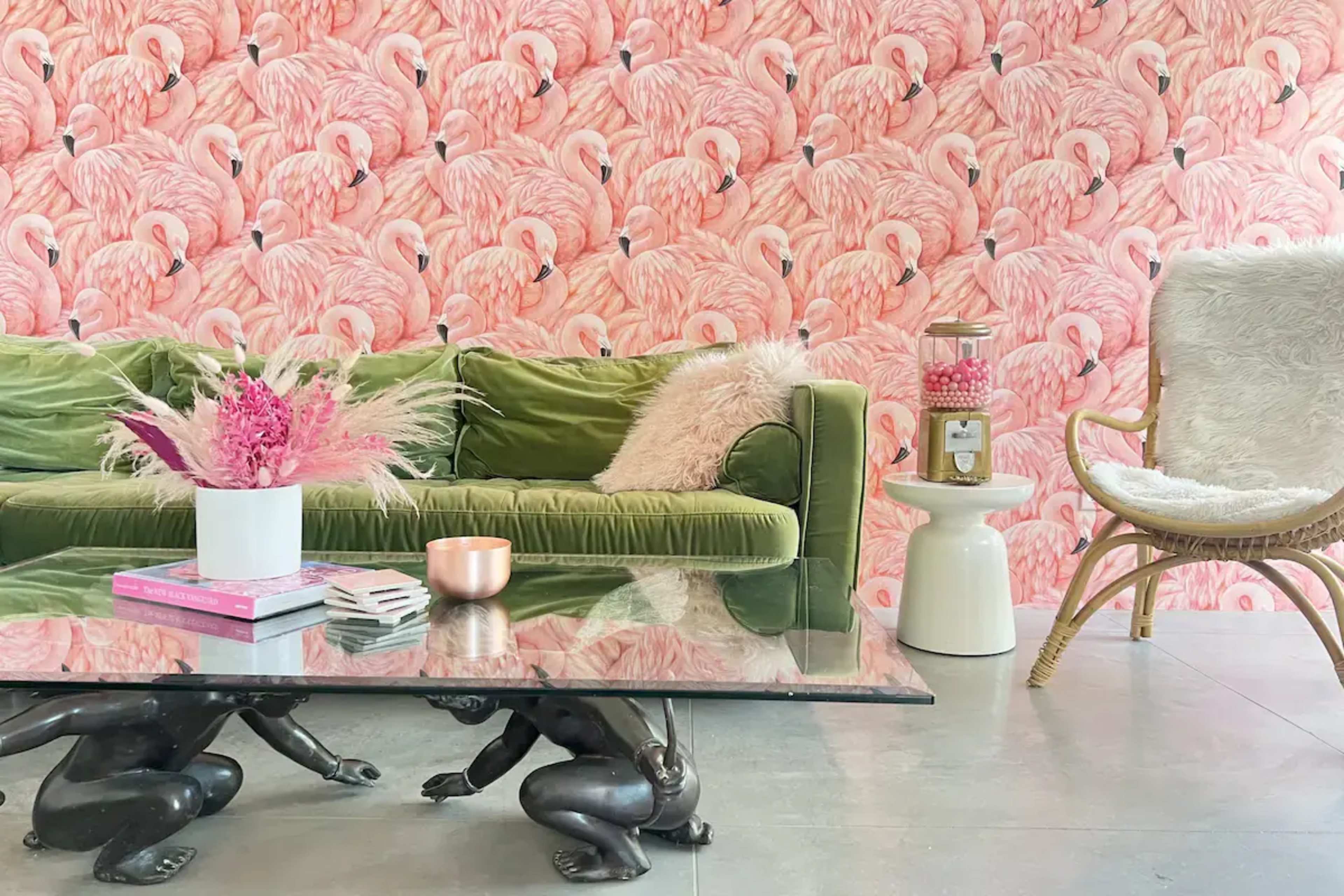 A green velvet sofa is positioned in front of a pink flamingo-patterned wallpaper, complemented by a glass coffee table and a rattan chair with a decorative cushion.