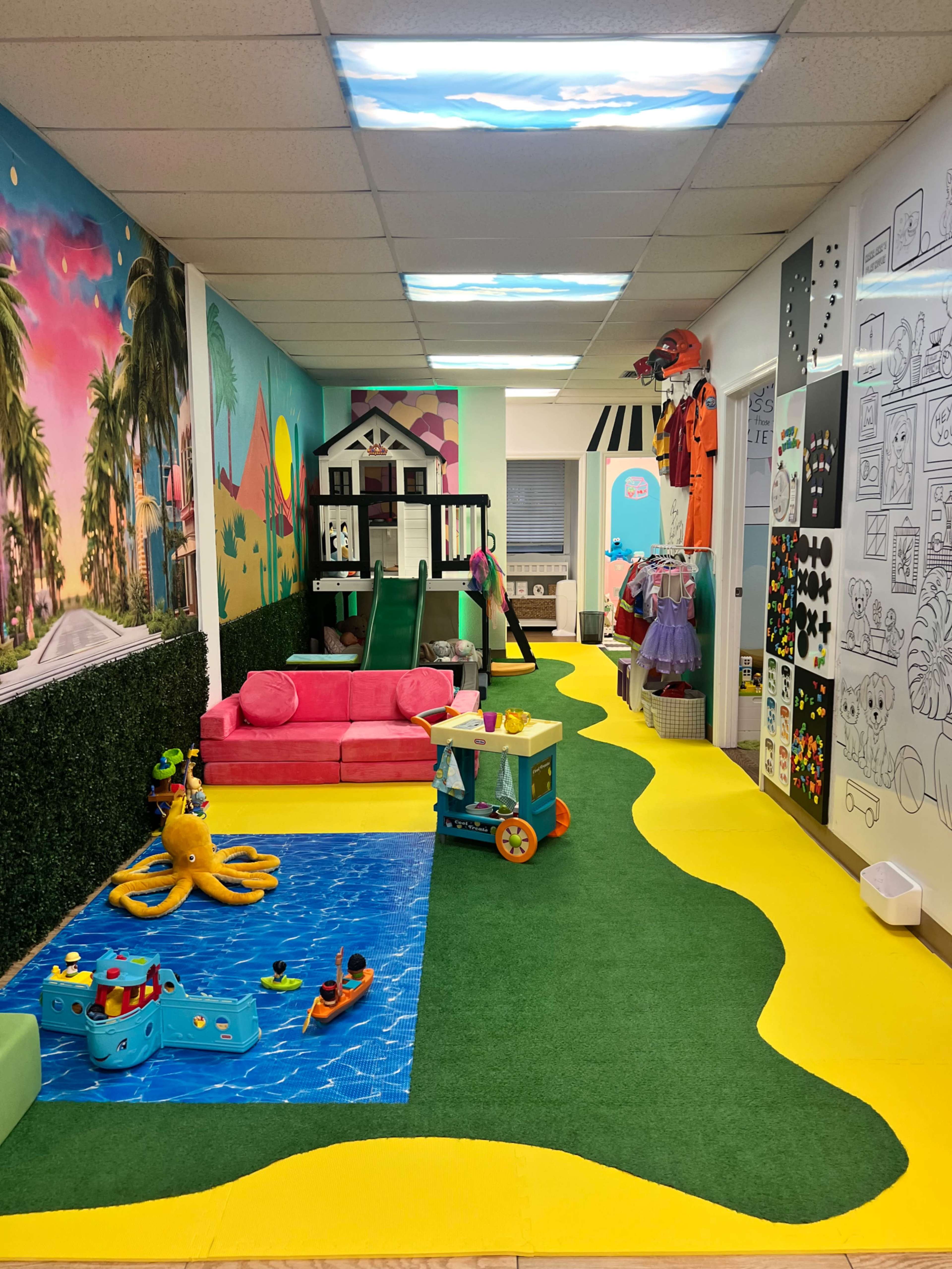 The image shows a colorful indoor play area featuring a green and yellow carpet, a water-themed play section, toys, and a small climbing structure.