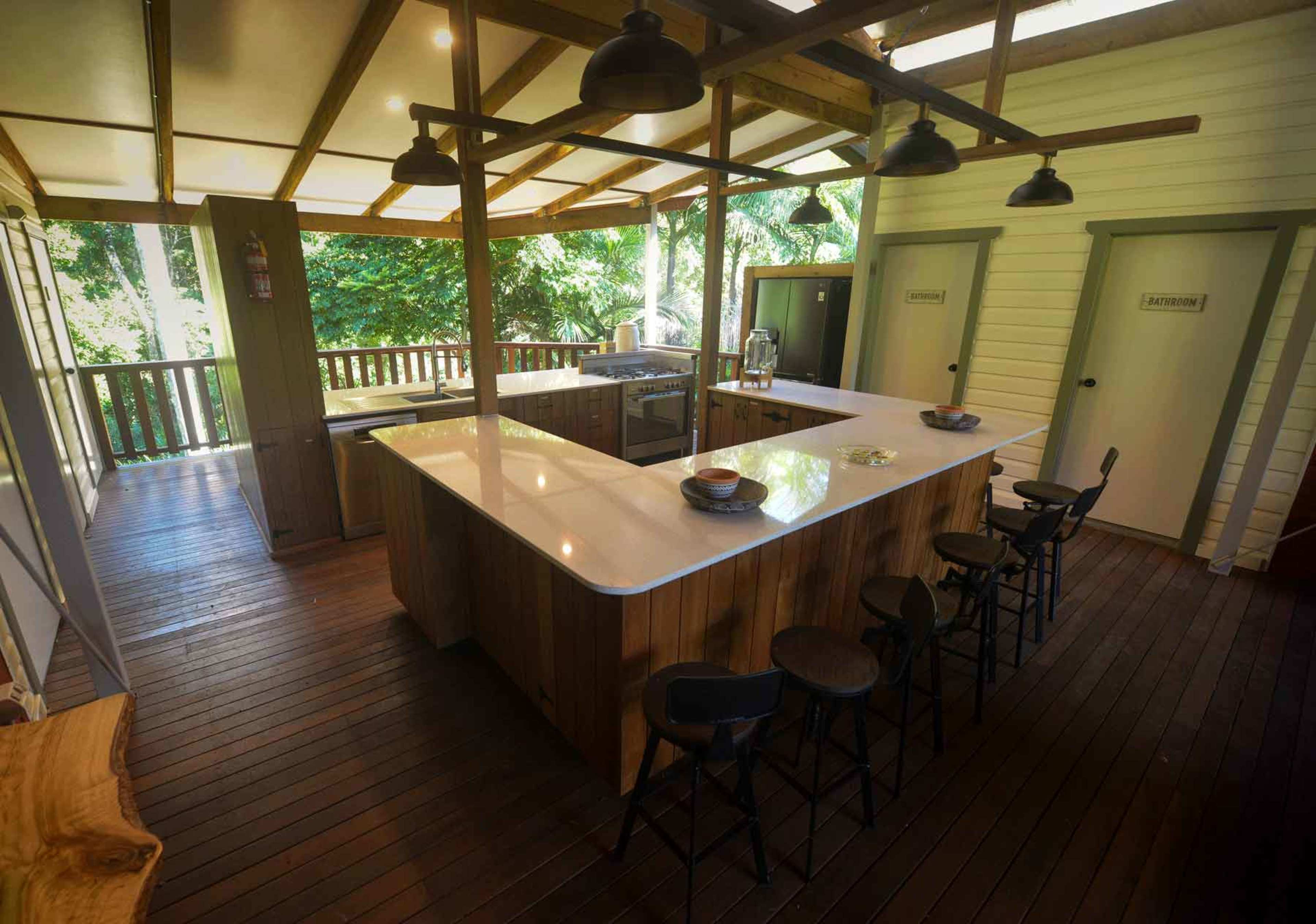 The image shows a modern kitchen with a large island and several bar stools, surrounded by wooden flooring and large windows overlooking greenery.