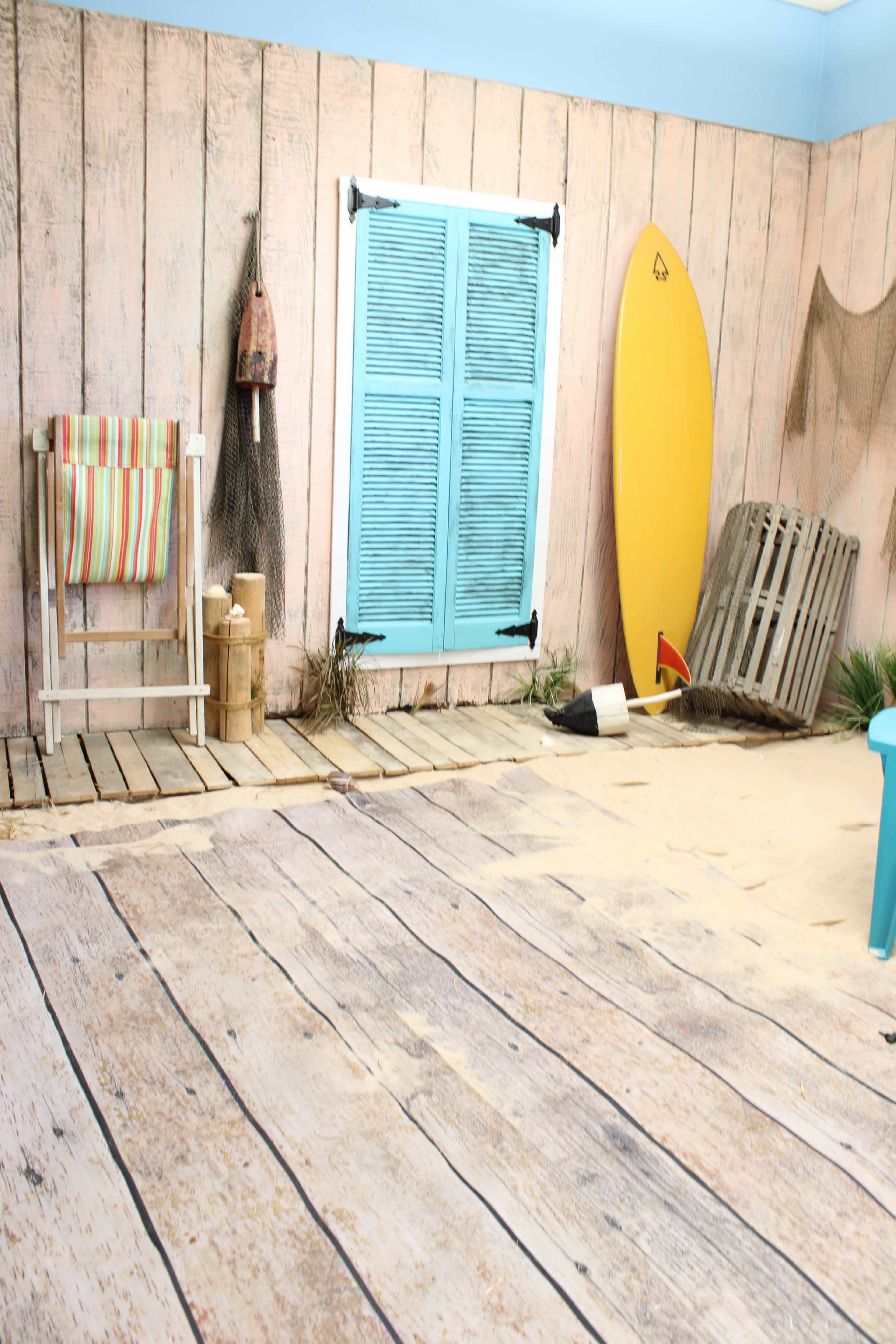 The image shows a beach-themed room with wooden walls, a sand-covered floor, a yellow surfboard, and a chair next to a window with blue shutters.