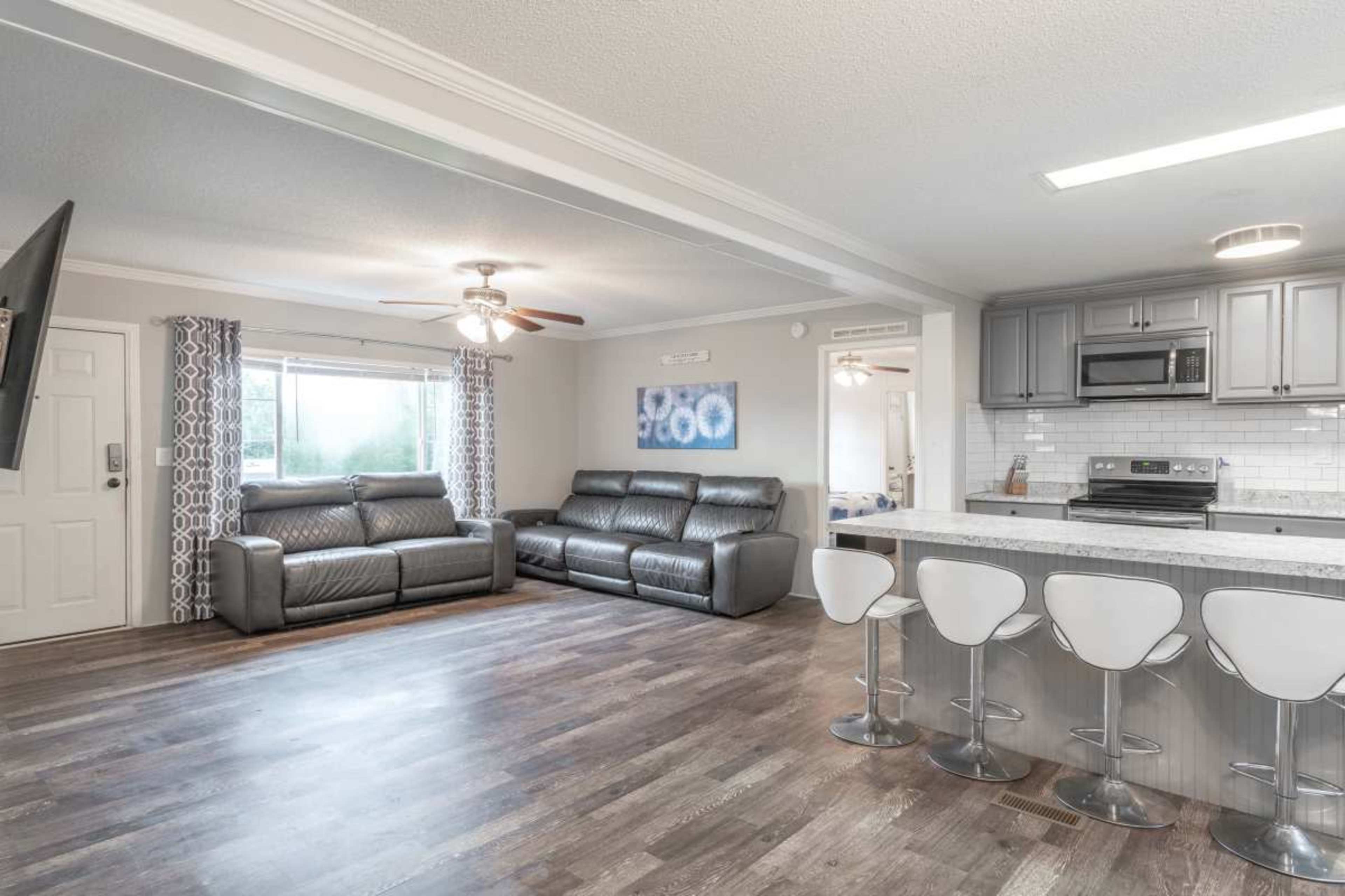 The image shows a modern living area with a kitchen bar, featuring gray leather sofas, a white countertop, and light-colored cabinetry.
