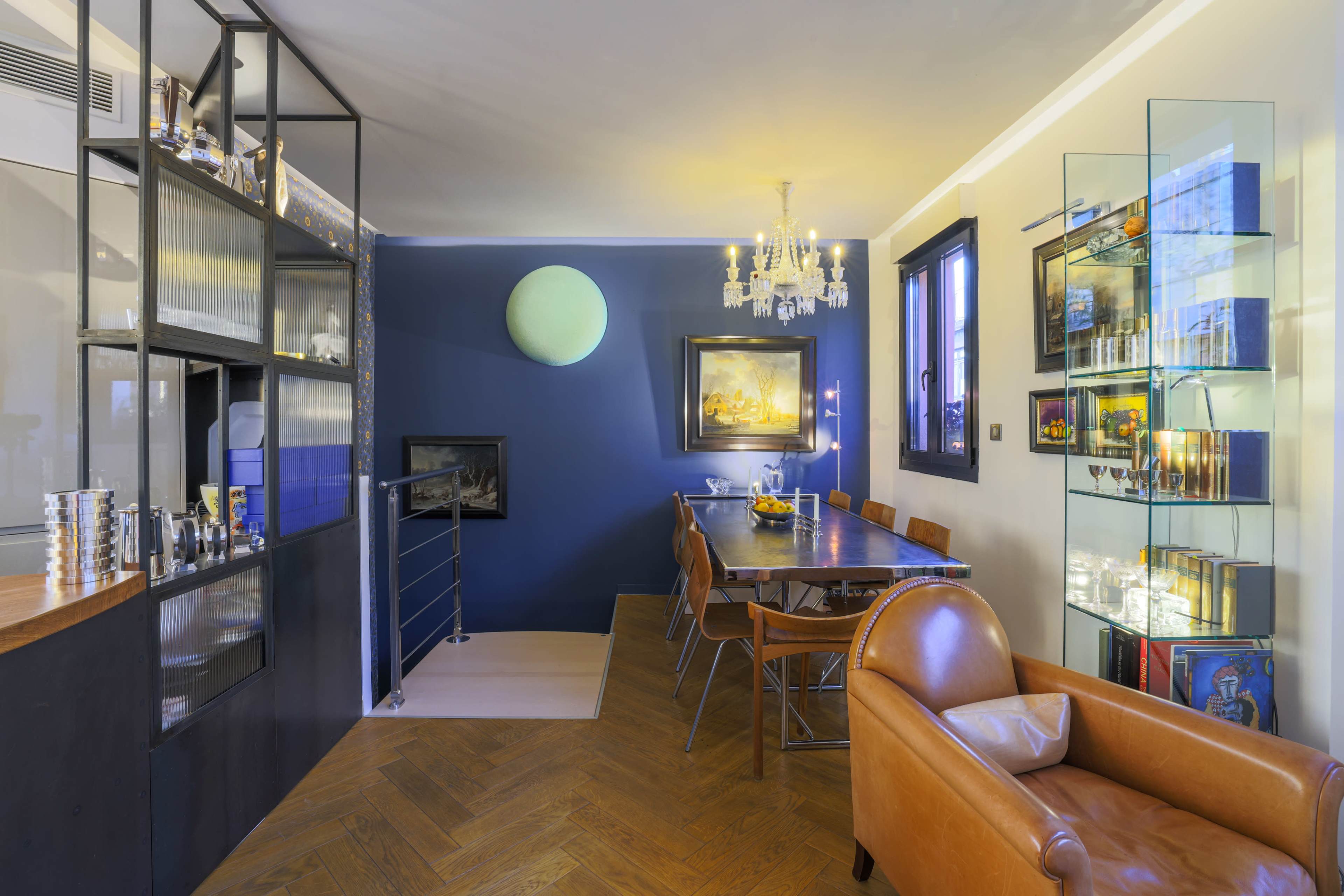 The image shows a modern dining area featuring a long table with wooden chairs, a glass display unit, and a vibrant blue wall with a round wall decoration.