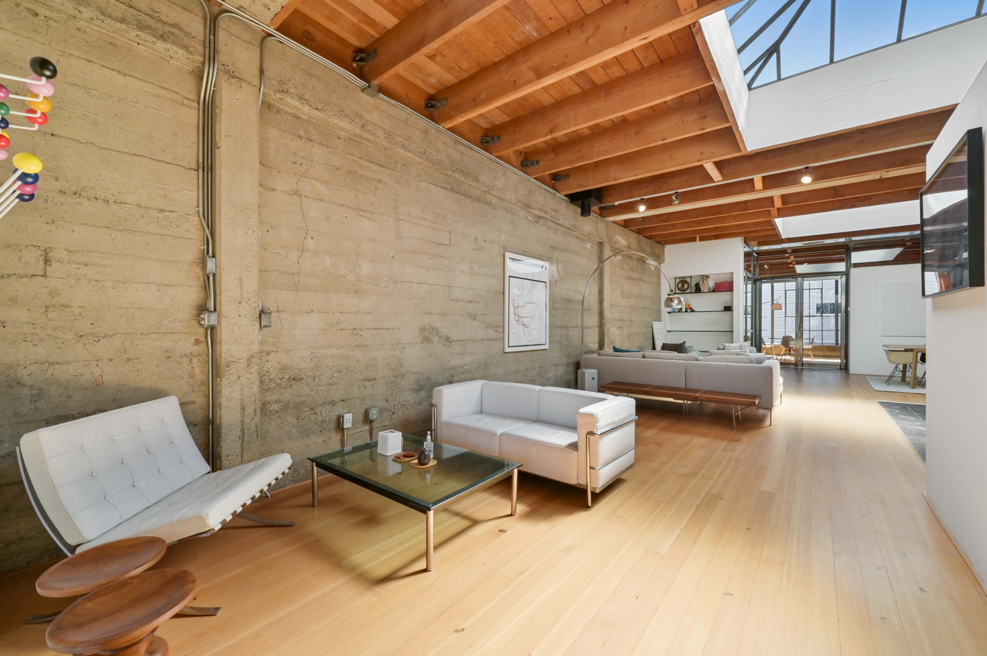 A spacious modern living area with concrete walls, wooden beams, and minimalist furniture arranged along a light hardwood floor.