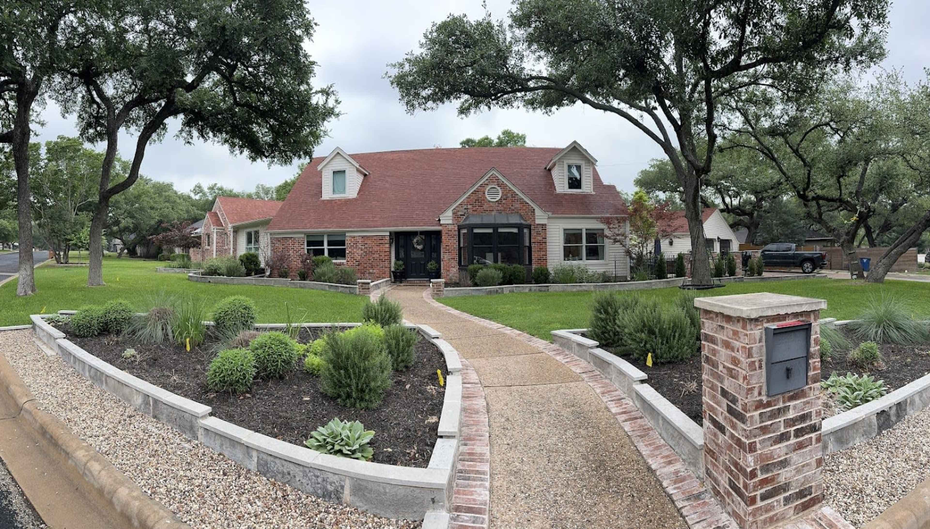 A brick house with a red roof and well-maintained landscaping is situated on a tree-lined street.