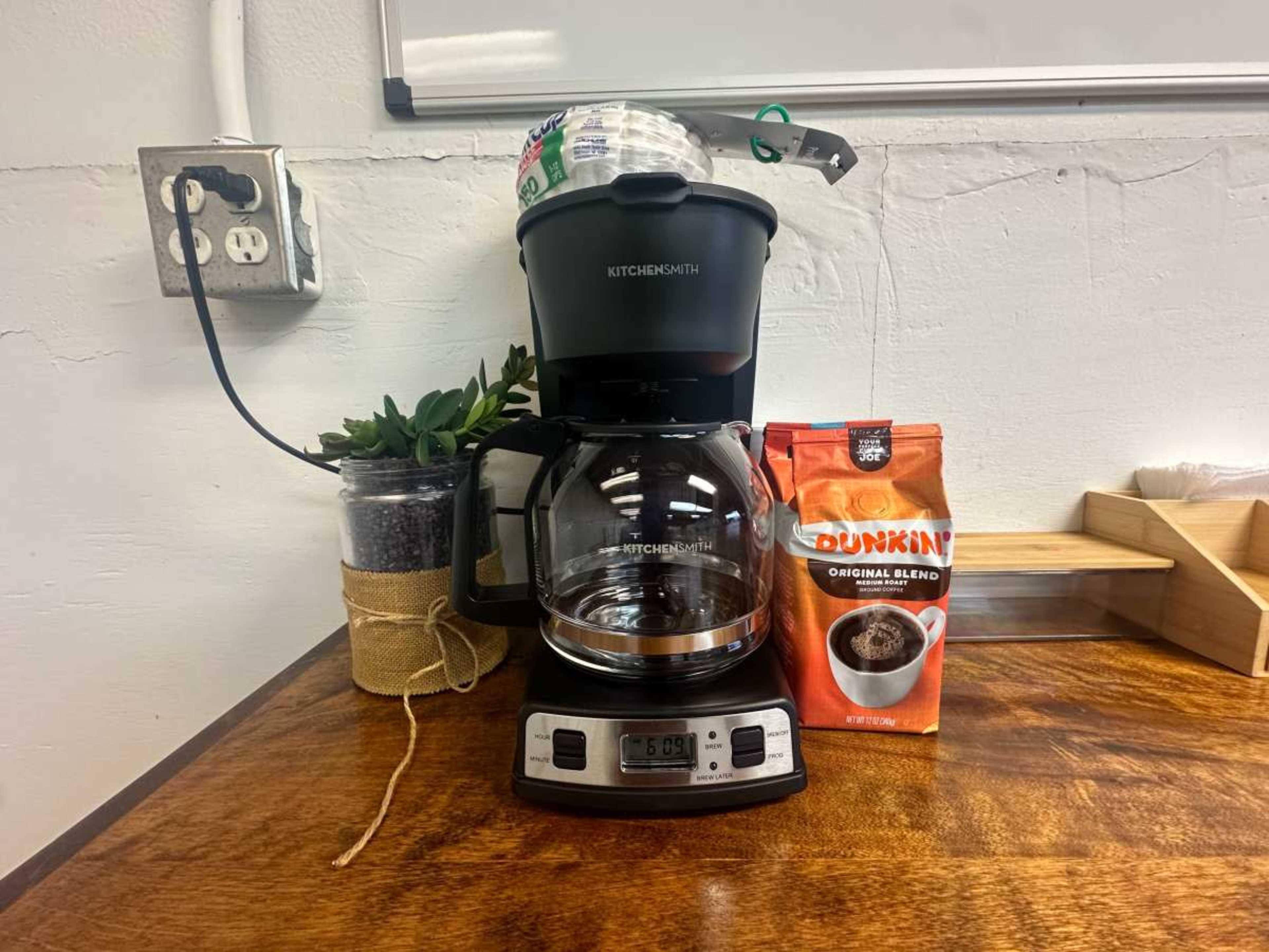 A coffee maker sits on a wooden counter next to a bag of Dunkin' coffee and a potted plant.