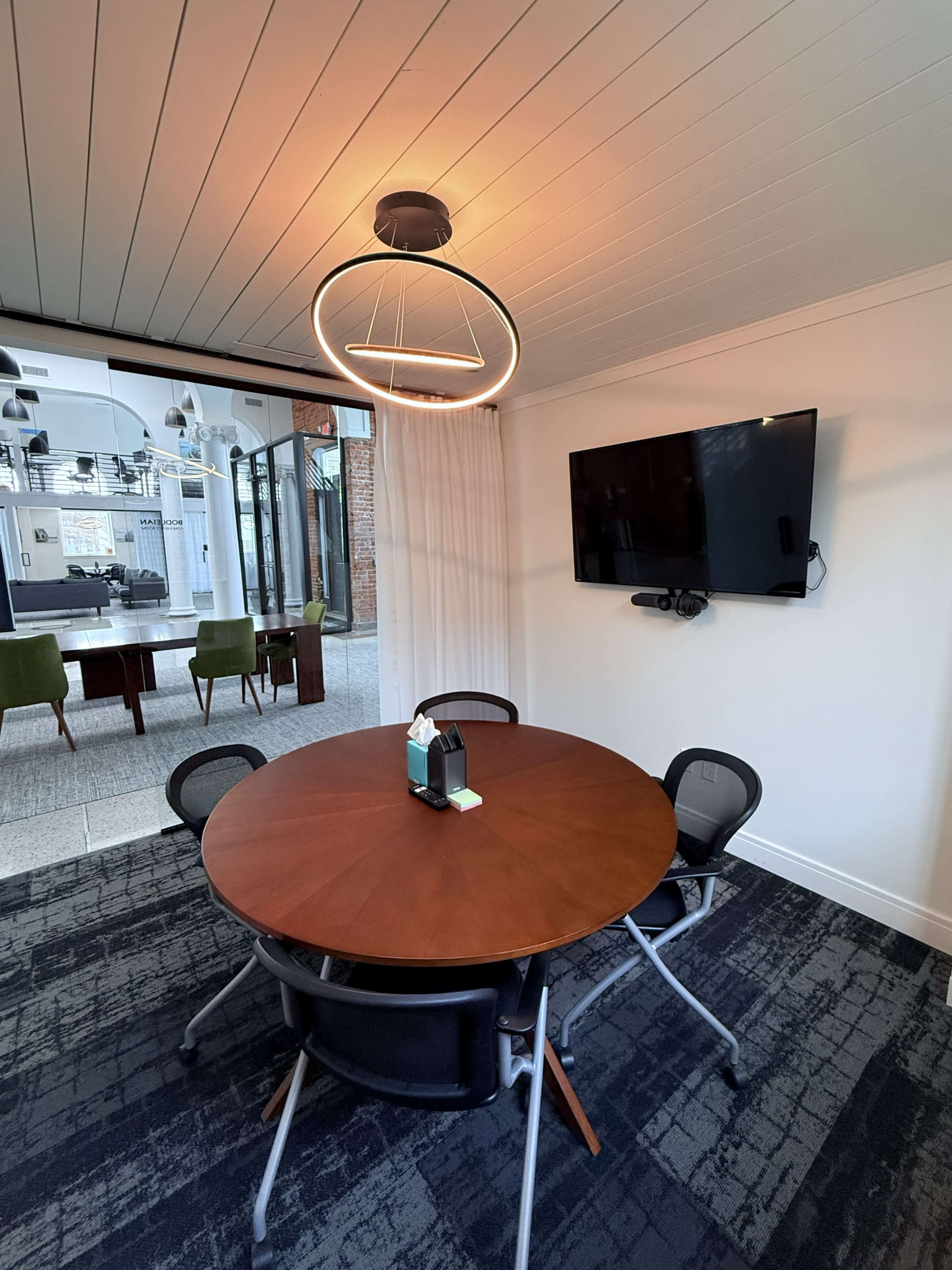 The image shows a modern conference room with a round wooden table, black chairs, a ceiling light fixture, and a wall-mounted TV.