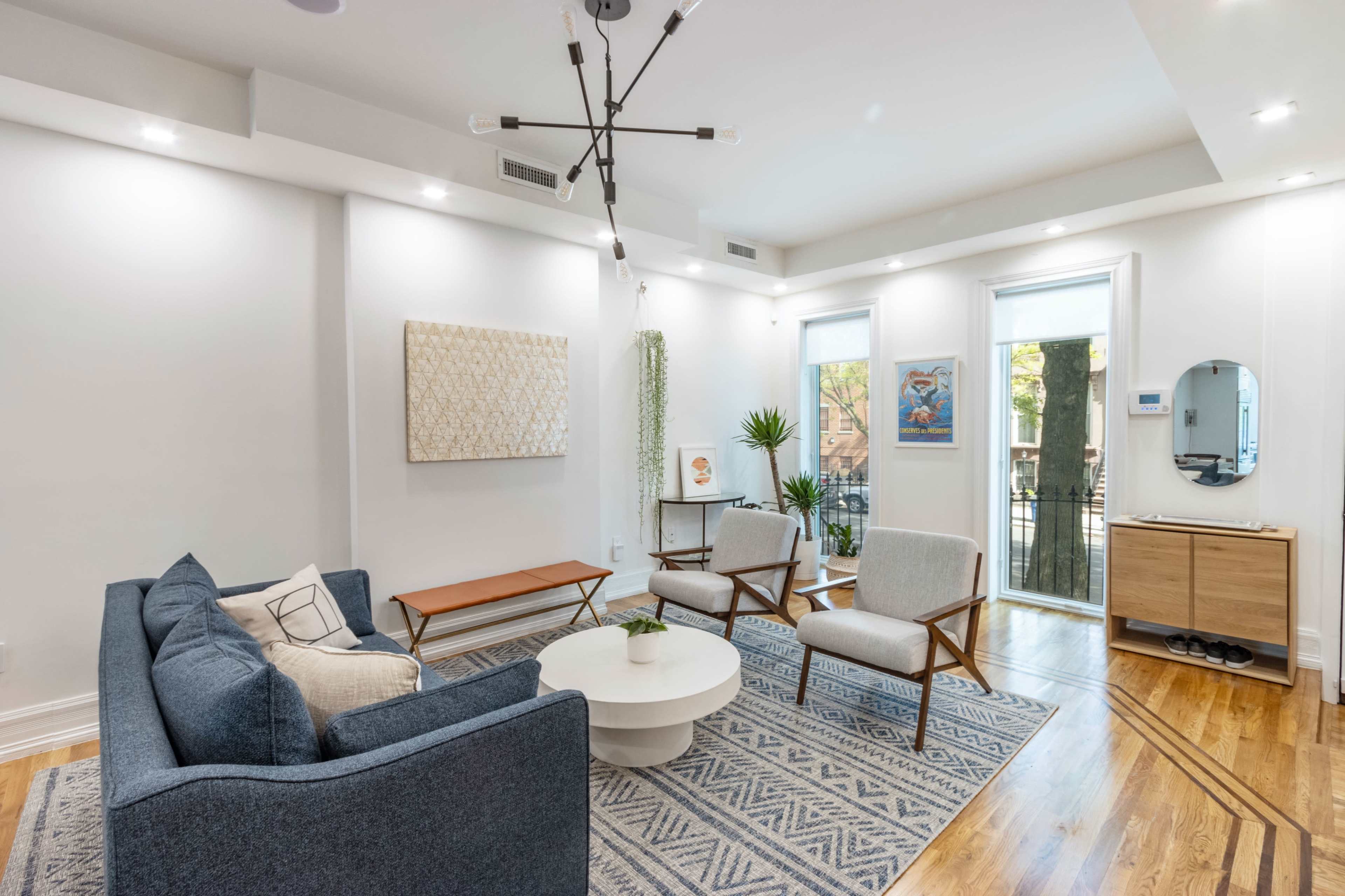 The image shows a modern living room with a blue sofa, two light chairs, a round coffee table, and large windows allowing natural light.